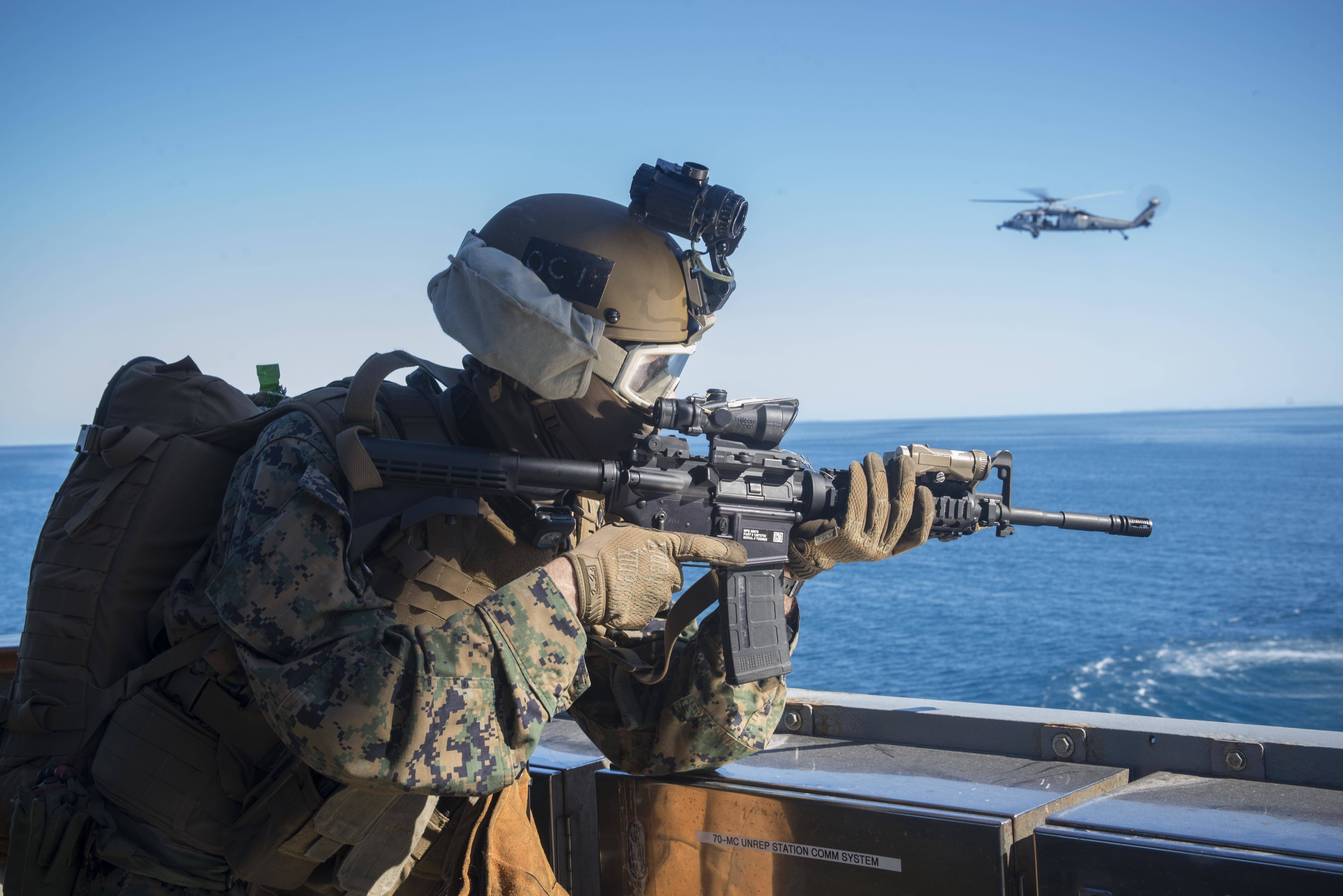海軍陸戰隊戰艦 壁紙 — 一名身穿迷彩服的士兵在船甲板上瞄準步槍，一架直升機在清澈藍天下的海洋上空飛行。