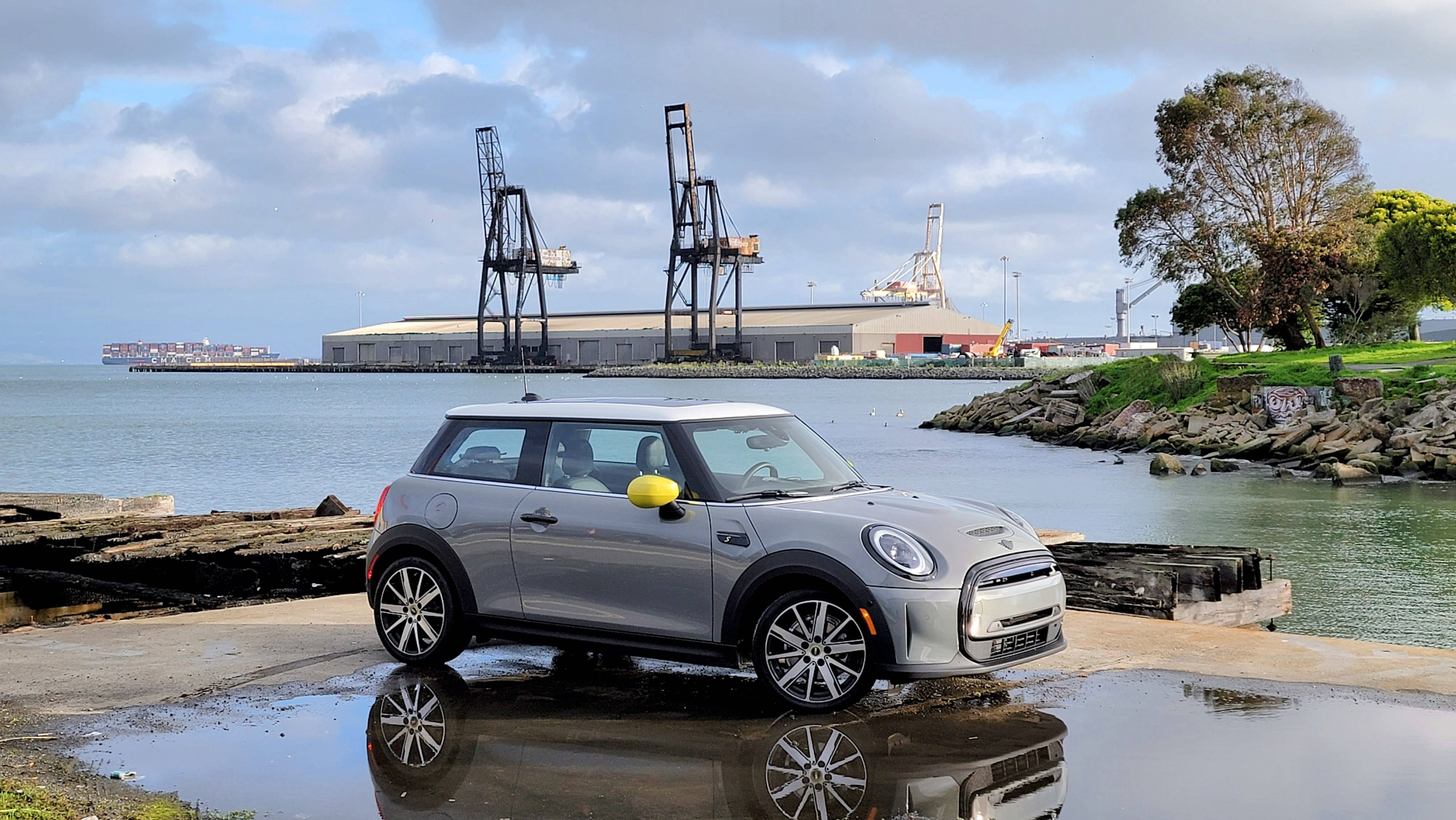 海邊停泊的白色迷你汽車 壁紙 — 一架銀色MINI Cooper停在濕滑的碼頭上，在水坑中倒映，背後有工業起重機和寧靜的水面。