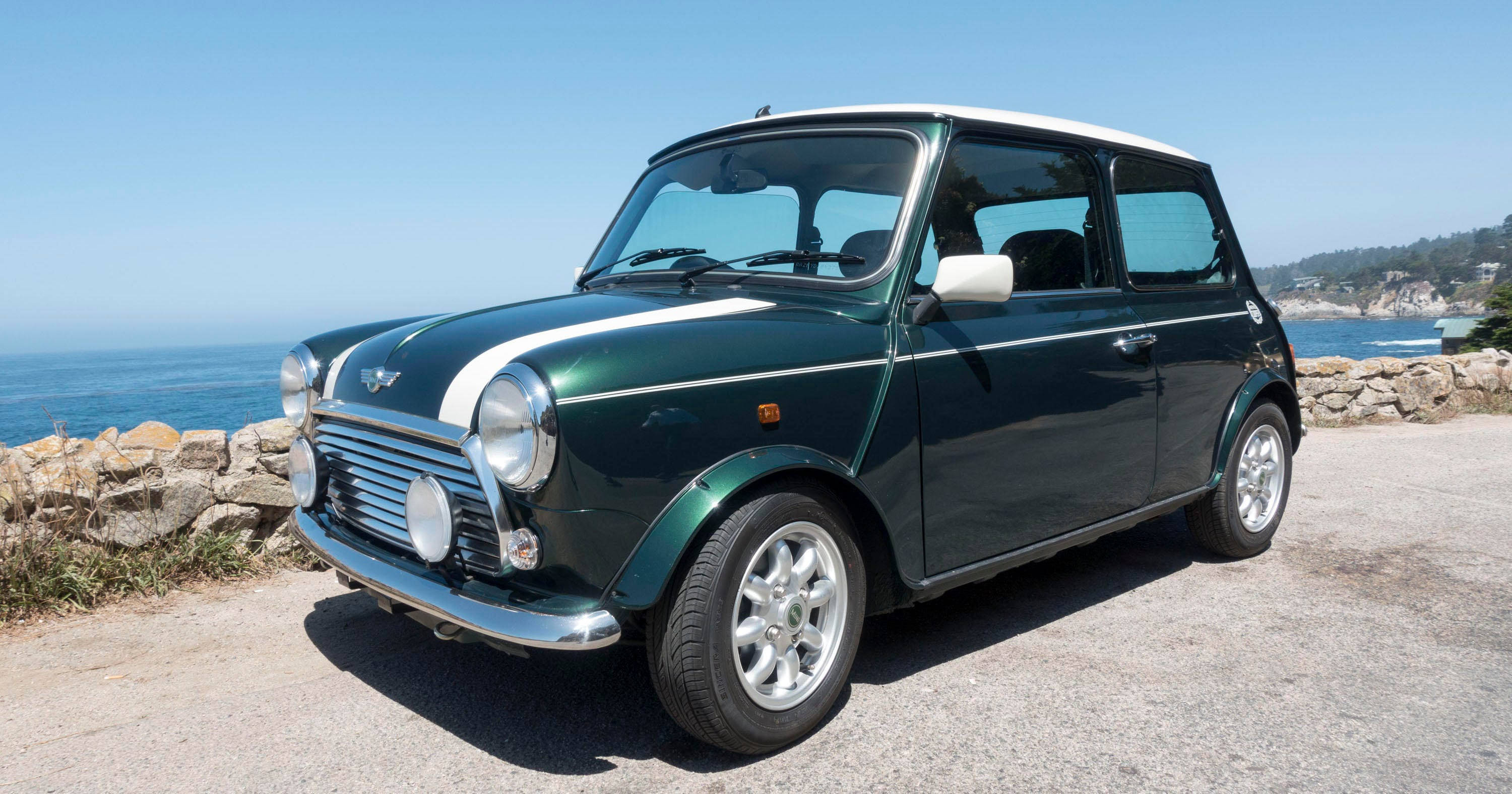 海邊停泊的綠白迷你車 壁紙 — 一輛經典的綠色Mini Cooper，白色車頂和賽車條紋，停在沿海公路上，俯瞰海洋，天空清澈湛藍。