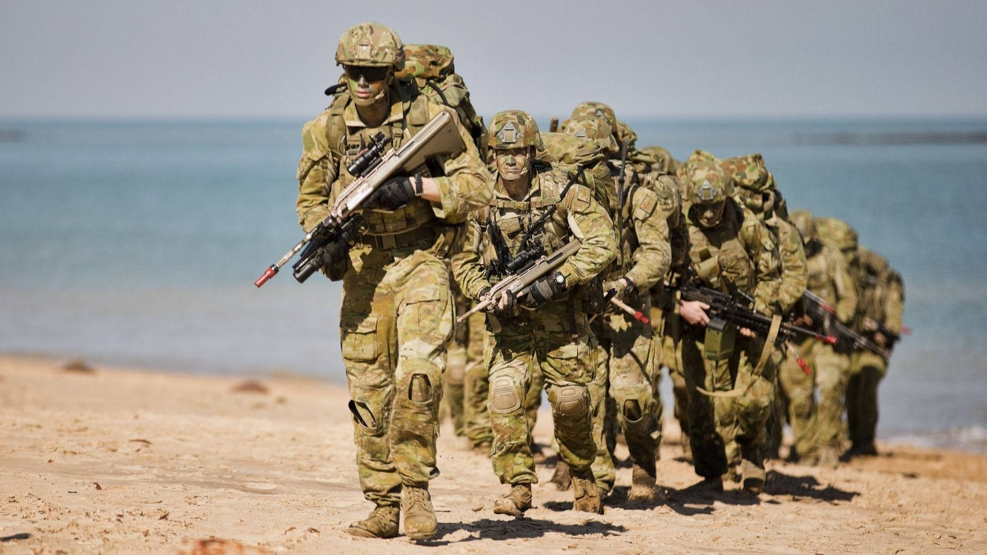 海邊列隊的軍人 壁紙 — 身穿迷彩裝備的士兵在陽光明媚的海灘上列隊行進，身後是延伸的海洋。