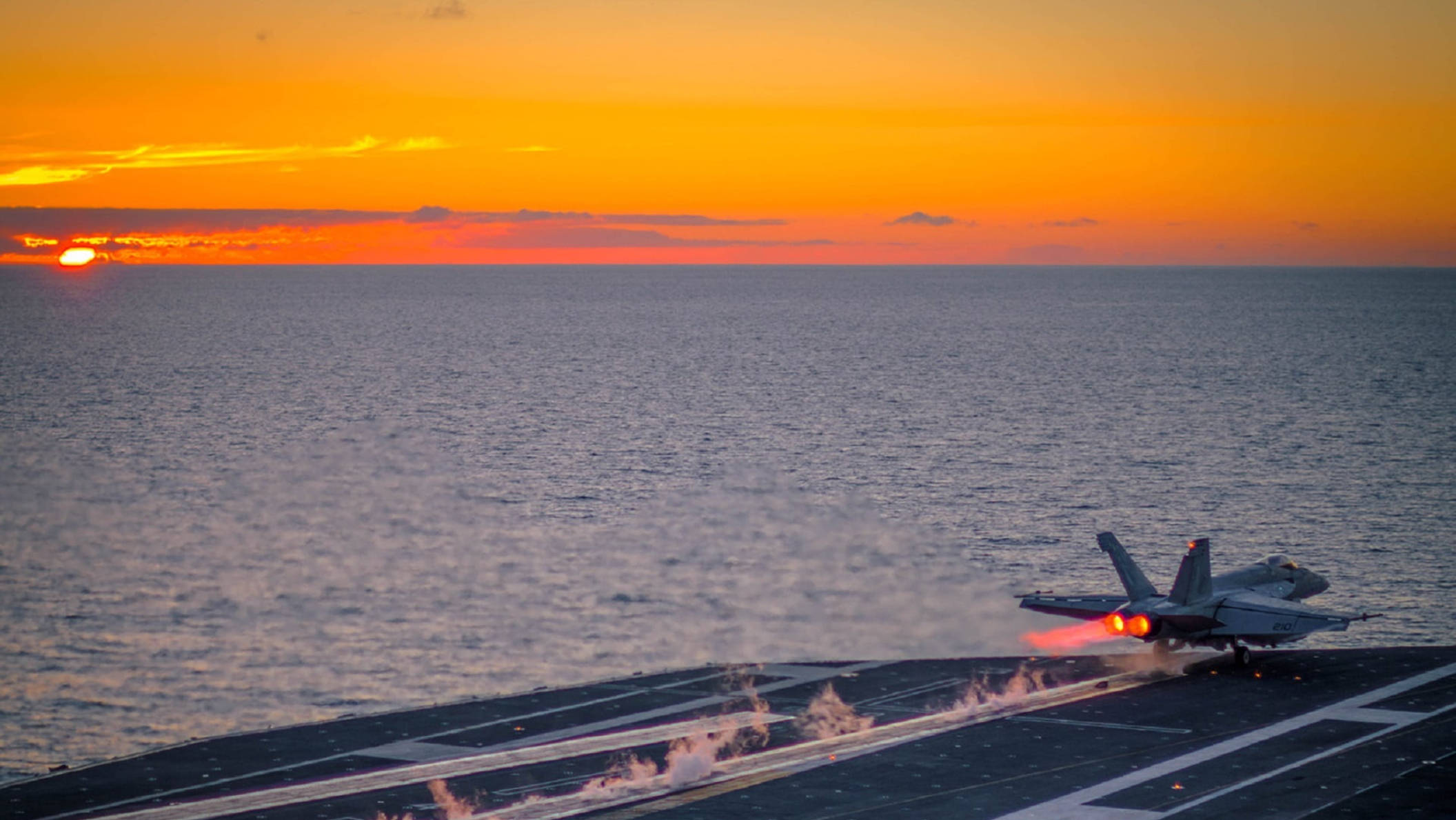 海邊跑道 壁紙 — 一架戰鬥機從航空母艦上起飛，夕陽在海平線上投下熾熱的橙色光芒。