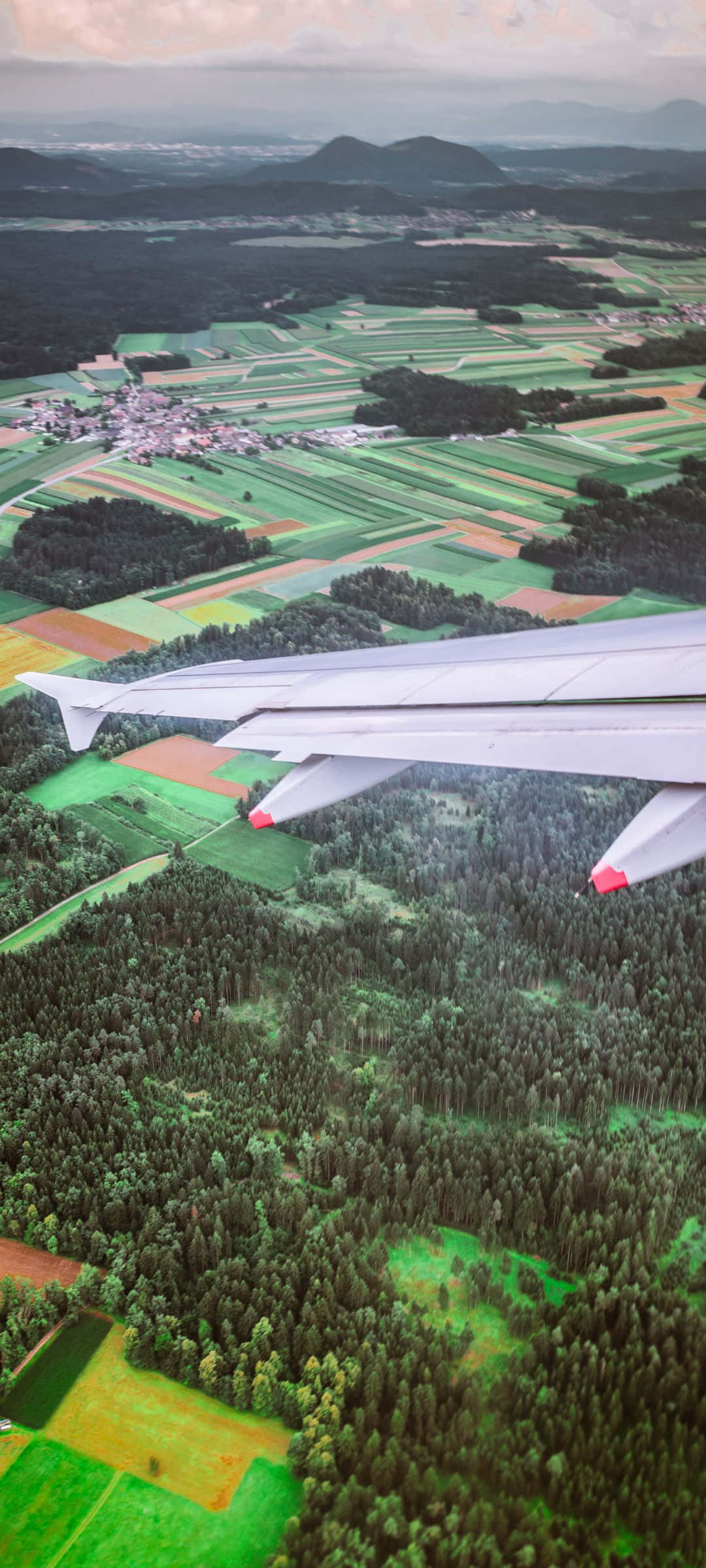 海面上寧靜的粉紅與藍色雲朵 壁紙 — 從飛機窗戶俯瞰，機翼下方是拼布般的綠色田野、森林和遠山，天空多雲。
