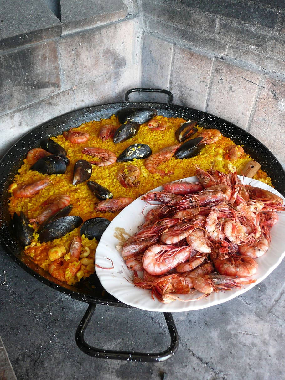 海鮮炒飯配生蝦 壁紙 — 一個大型西班牙海鮮飯鍋裝滿金黃色米飯和海鮮，旁邊放著一盤新鮮粉紅蝦，背景是灰色磚牆。