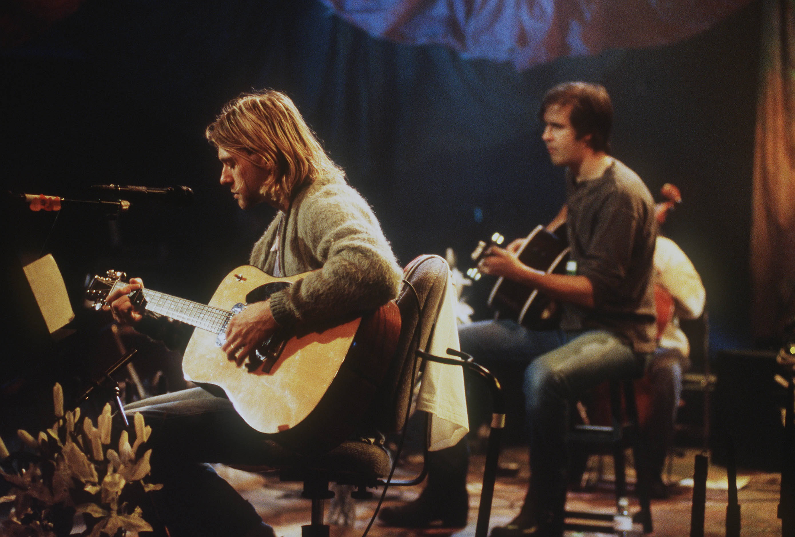 涅槃樂隊 4K MTV Unplugged 壁紙 — 一位長髮音樂家在燈光昏暗的舞台上彈奏木結他，另一位樂隊成員在溫暖的聚光燈下在他旁邊彈奏。