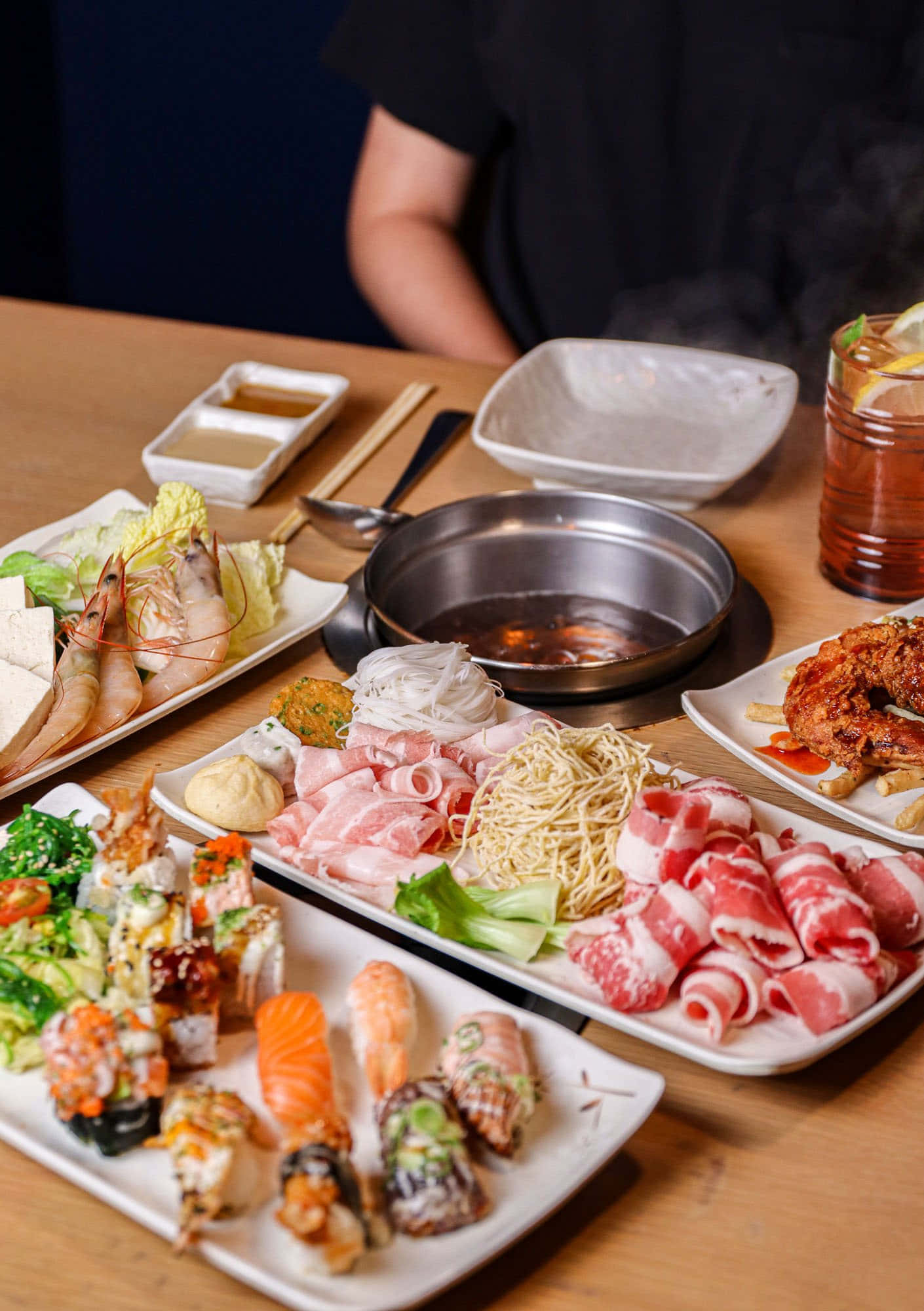 涮涮鍋食材盤：肉與蔬菜 壁紙 — 一個冒煙的熱鍋，周圍放著壽司、刺身、生牛肉卷和蝦的碟子，在木桌上。