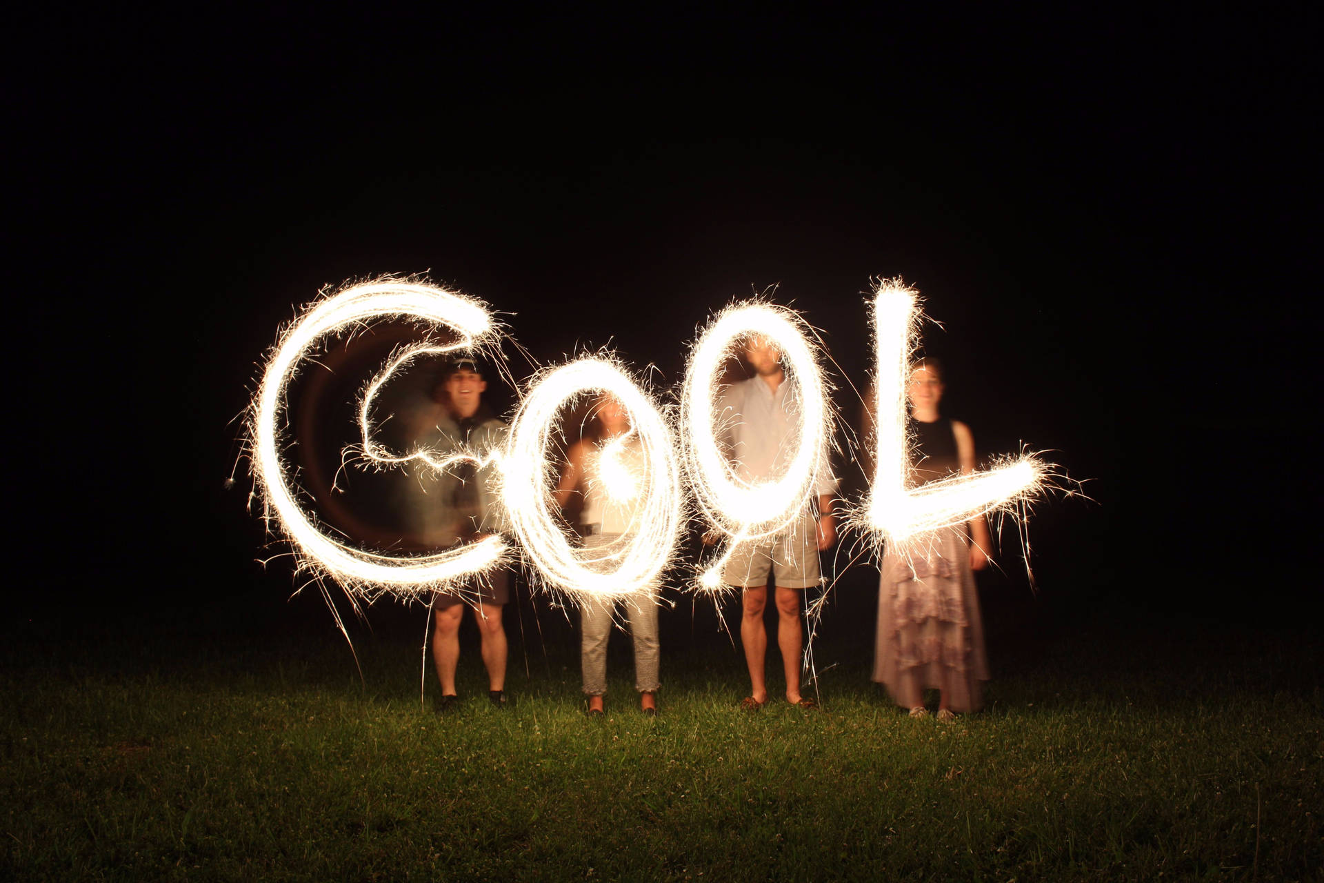 四個人在夜晚的草地上手持煙花棒，拼出COOL。