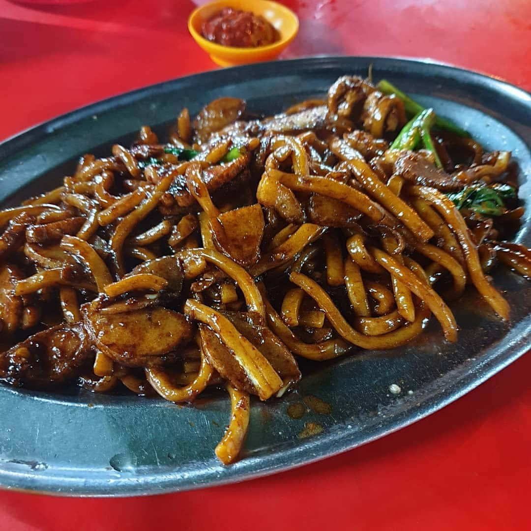淋上深色醬汁的馬來西亞福建麵 壁紙 — 一盤熱騰騰的炒麵，配上肉和青菜，在紅色桌子上閃閃發光。