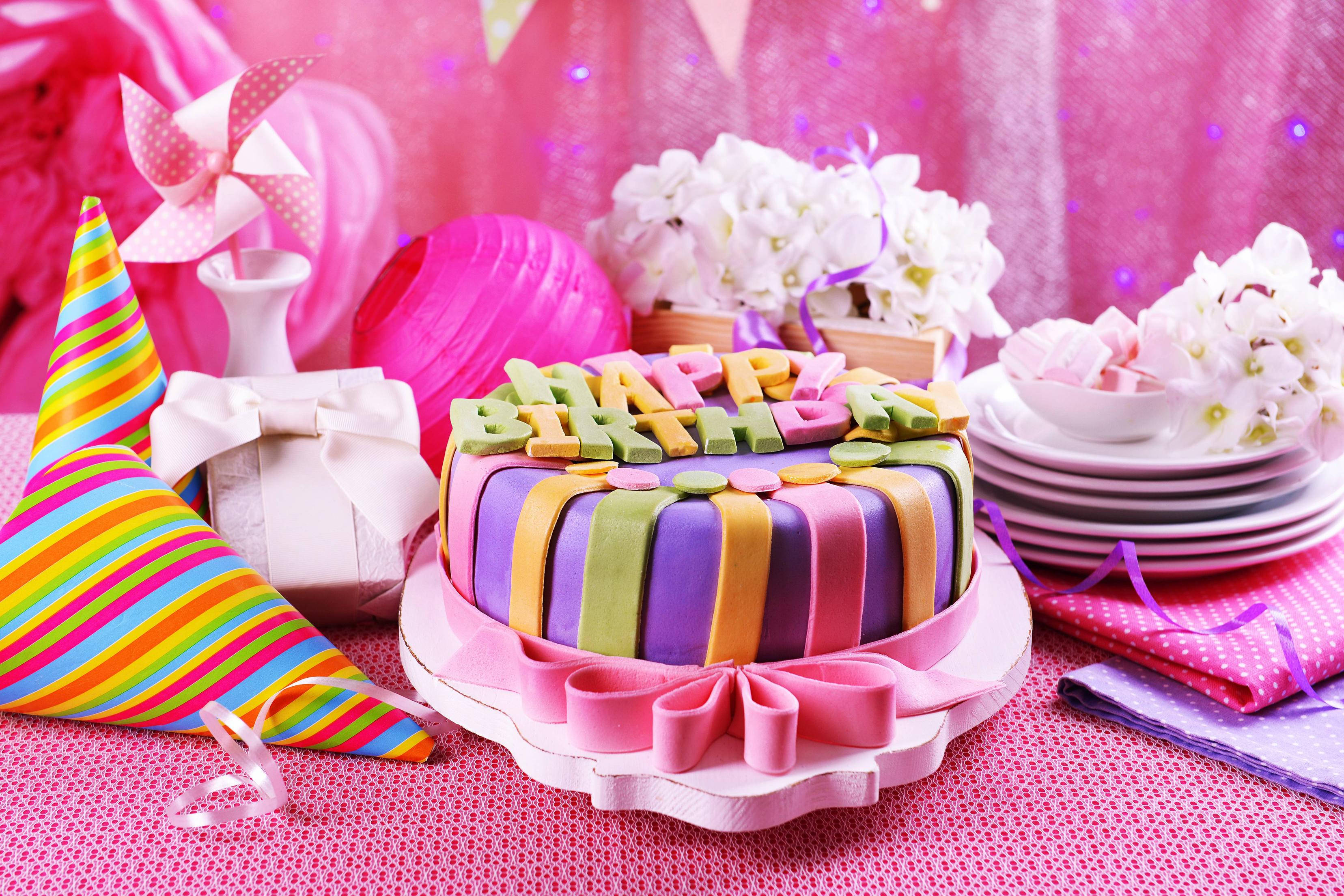淡彩生日蛋糕 壁紙 — 一個色彩繽紛的生日蛋糕，上面有彩色翻糖字母和絲帶，放在粉紅色桌布上，周圍環繞著派對帽和白色花朵。