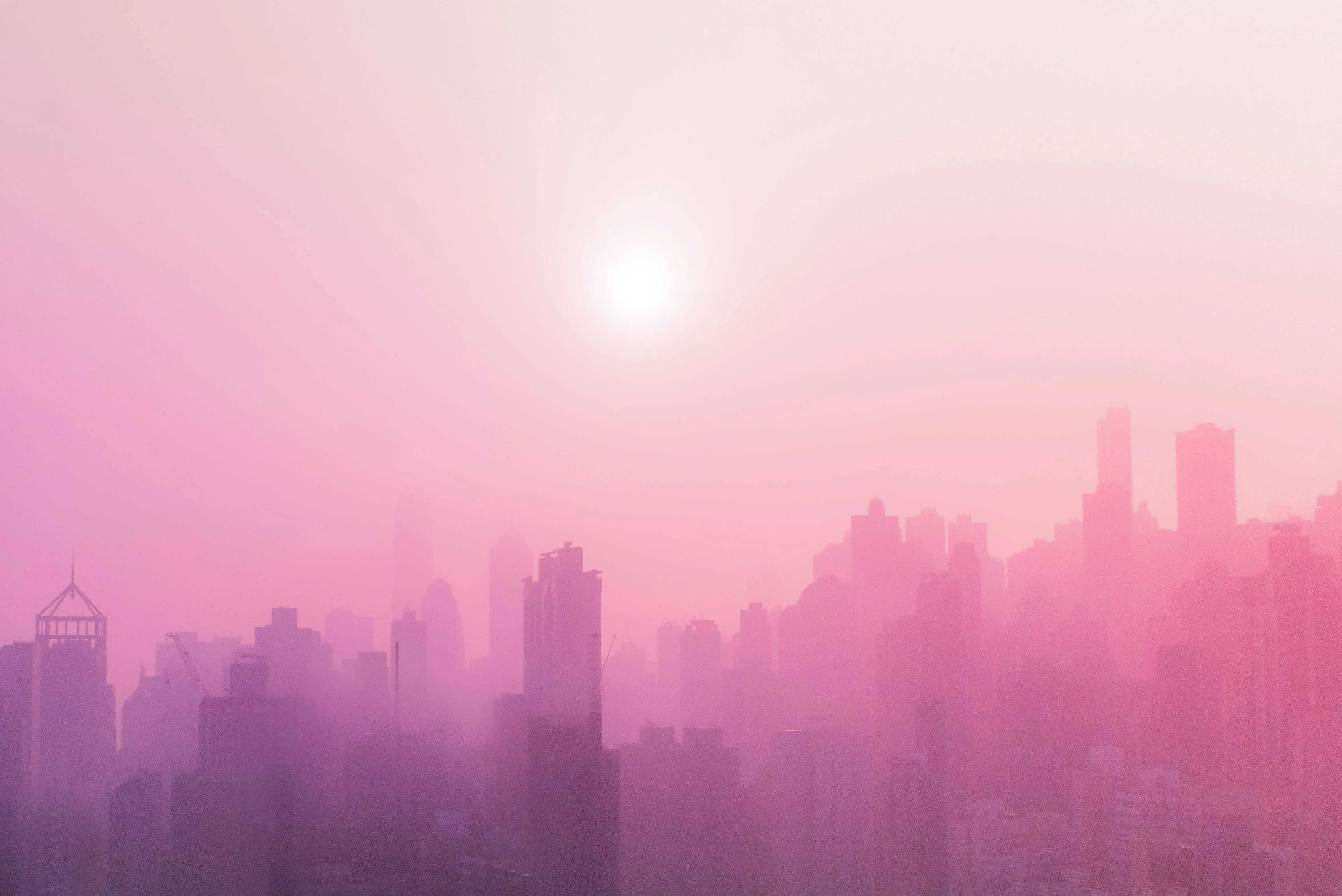 淡粉城市風景 壁紙 — 朦朧的城市天際線沐浴在柔和的粉紅色和紫色色調中，太陽在頭頂明亮地照耀。