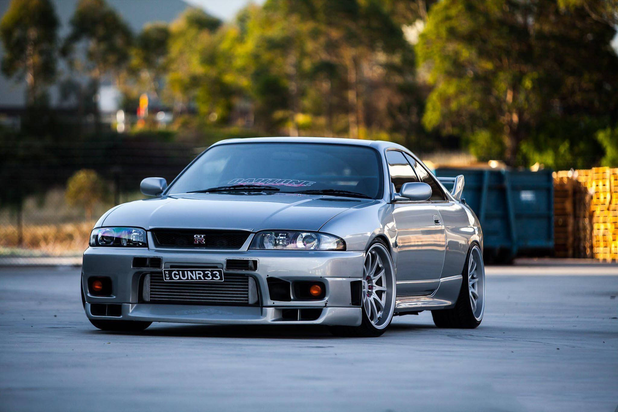 淡藍色Nissan Skyline GTR R33 壁紙 — 一輛銀色日產Skyline R34跑車，配備訂製輪圈和降低懸掛，停泊在混凝土上，背景有樹木和垃圾箱。