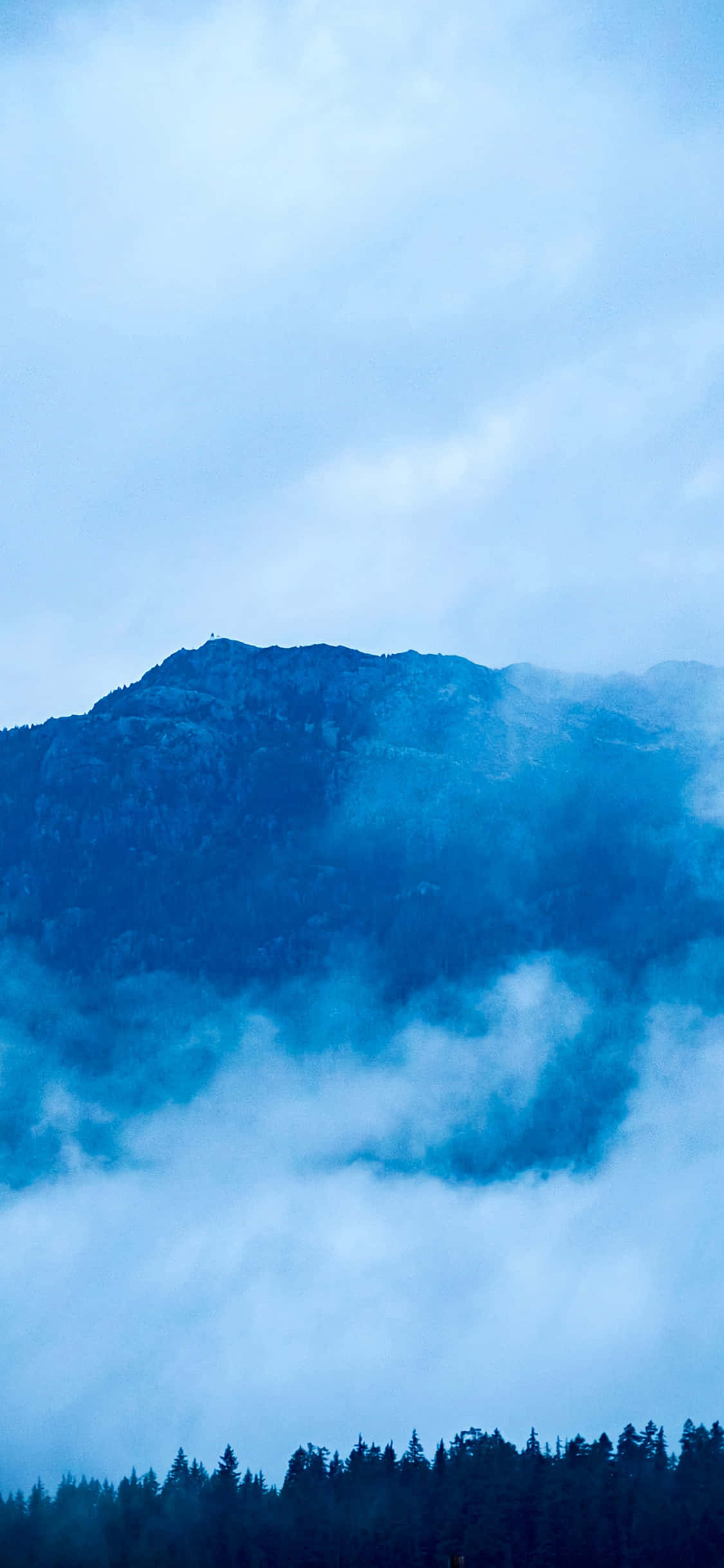 iPhone X Nature 桌布 壁紙 — 淡藍色天空下，煙霧繚繞的山峰從旋轉的雲層中浮現，底部有黑暗的森林輪廓。