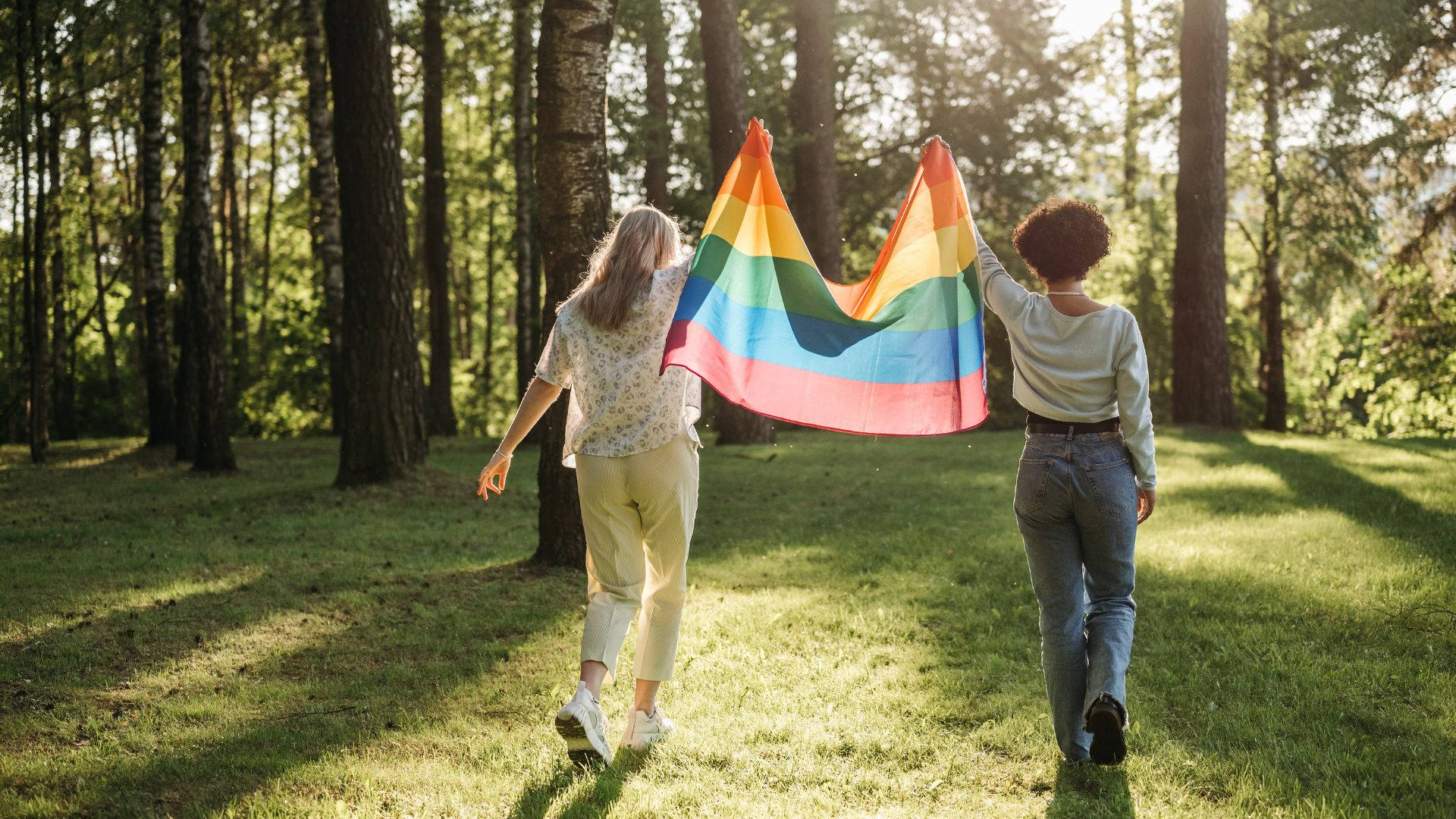 淡雅 LGBT 女性舉旗 壁紙 — 兩個人穿過陽光明媚的森林，高舉著鮮豔的彩虹旗。