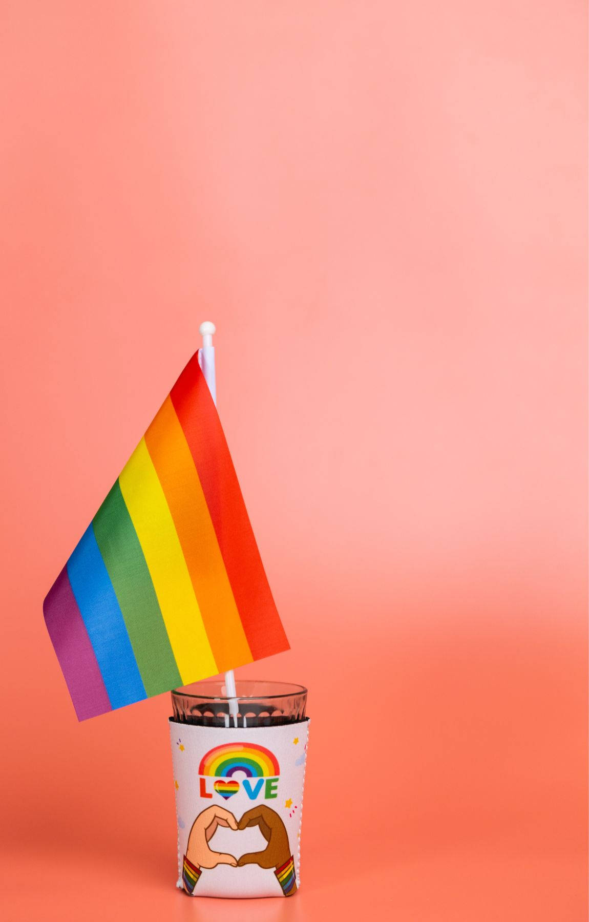 淡雅 LGBT 旗幟置於玻璃上 壁紙 — 一支彩虹旗從一個帶有彩虹心形和兩隻手形成心形符號的杯子中伸出，背景為純色蜜桃色。