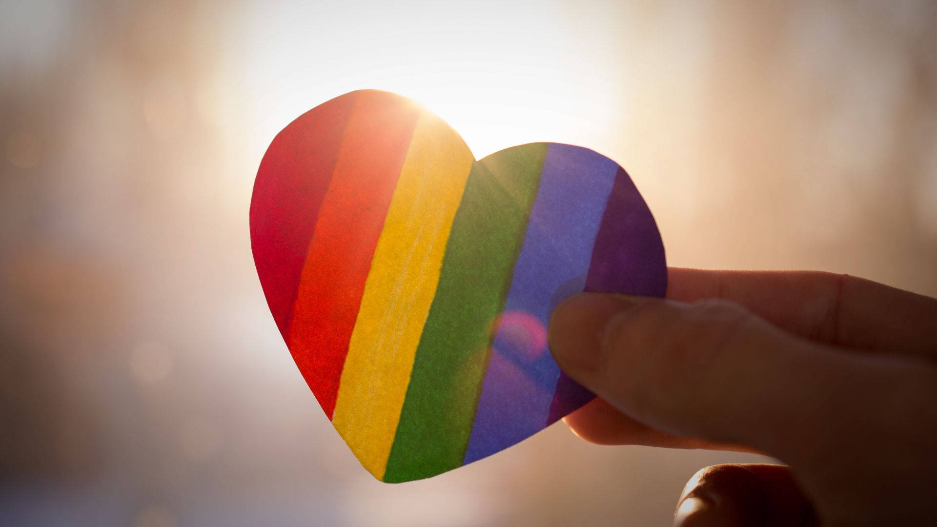 淡雅 LGBT 雨虹心形 壁紙 — 一隻手拿著彩虹色的心形剪紙，背景是明亮的陽光和柔和散景。