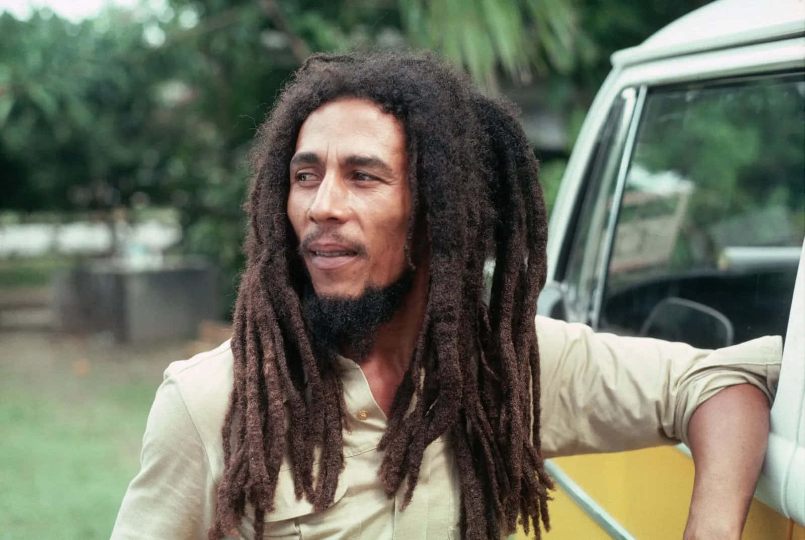 深受愛戴的牙買加雷鬼藝術家鮑勃·馬利 壁紙 — Bob Marley 靠在一輛復古貨車上，身後是茂密的綠色樹葉，一幅坦率的戶外肖像。