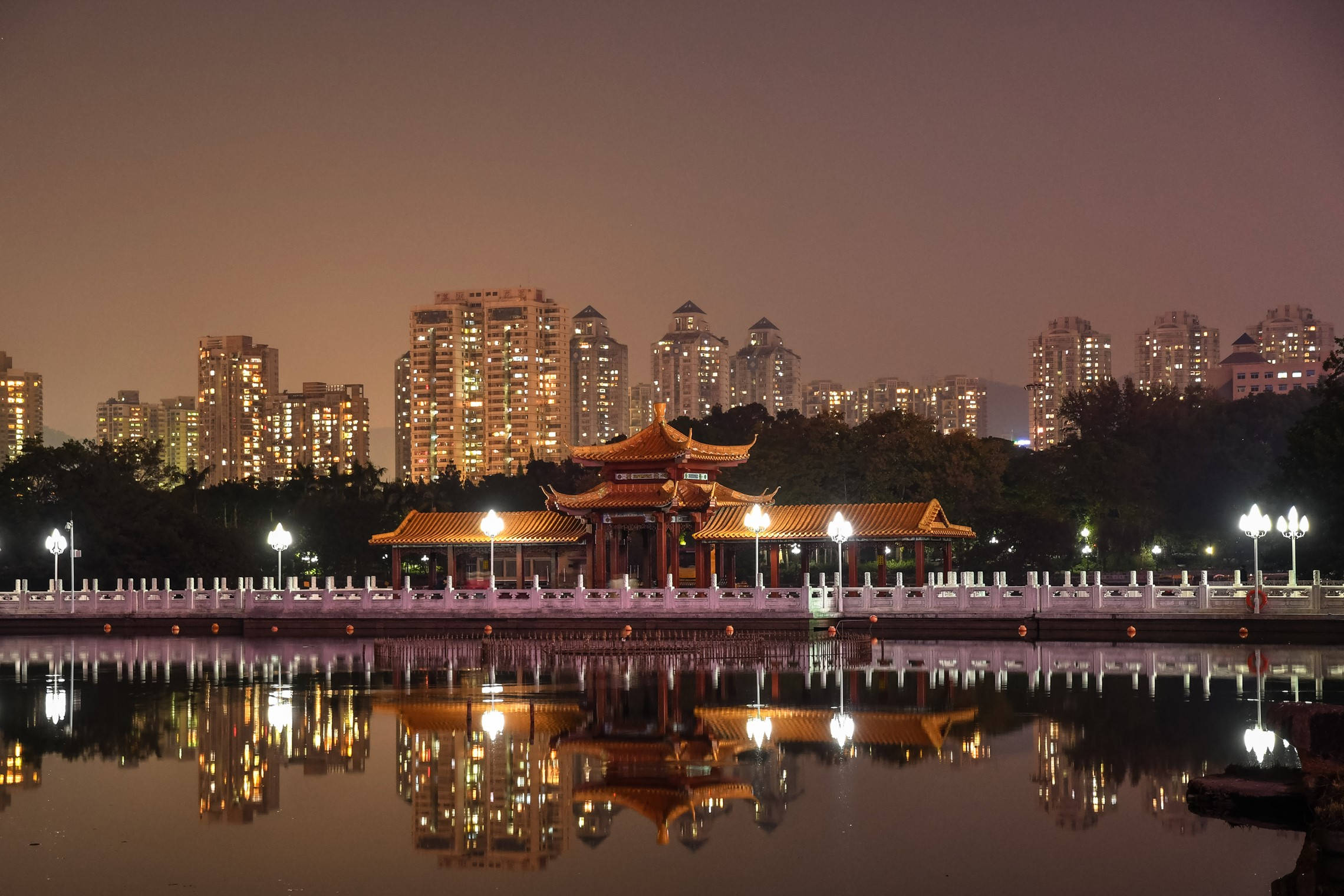 深圳傳統廟宇 壁紙 — 一座傳統中式亭閣在夜晚溫暖地發光，其倒影完美地映照在平靜的水面上，背景是燈火通明的城市摩天大樓。