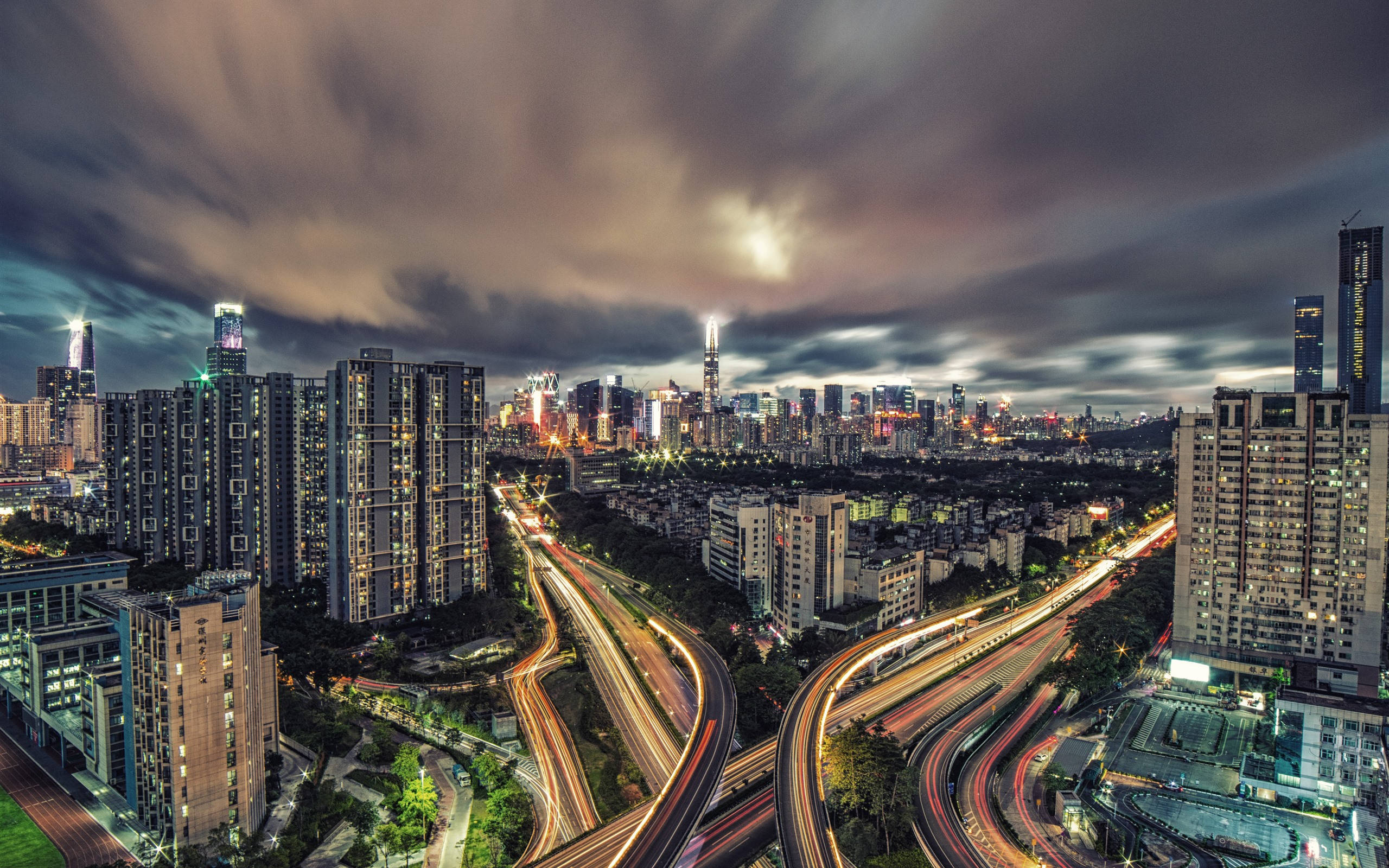 深圳夜間放鬆 壁紙 — 黃昏時分充滿活力的城市景觀，閃爍的摩天大樓和車流的光跡在戲劇性的雲層下。