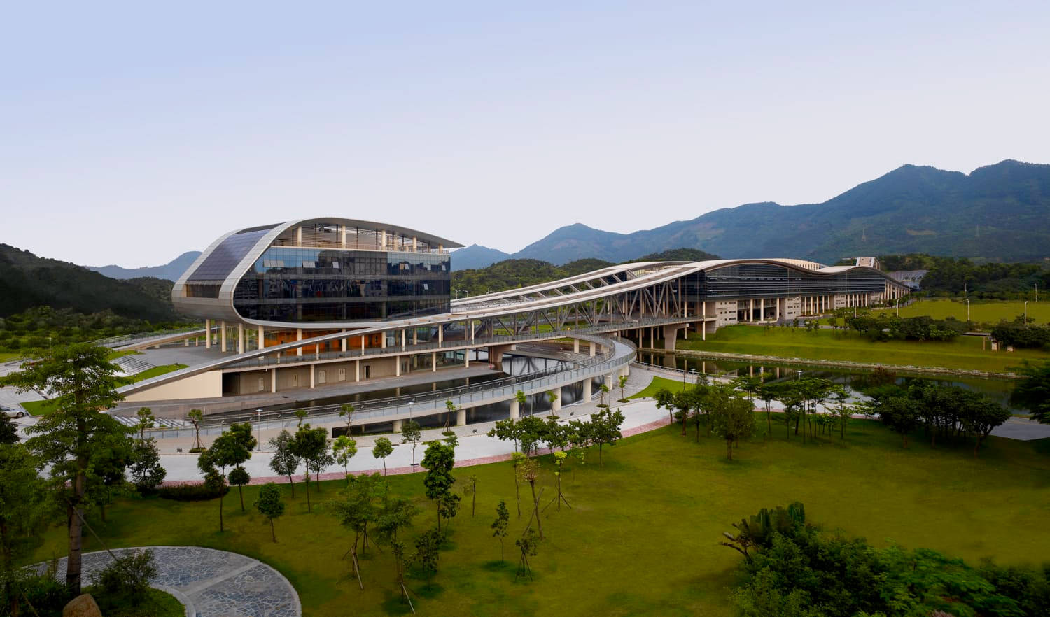 深圳大學 壁紙 — 一座未來風格的玻璃和鋼結構建築，採用弧形設計，坐落在茂密的綠色草坪和遠處山脈之間，天空晴朗。