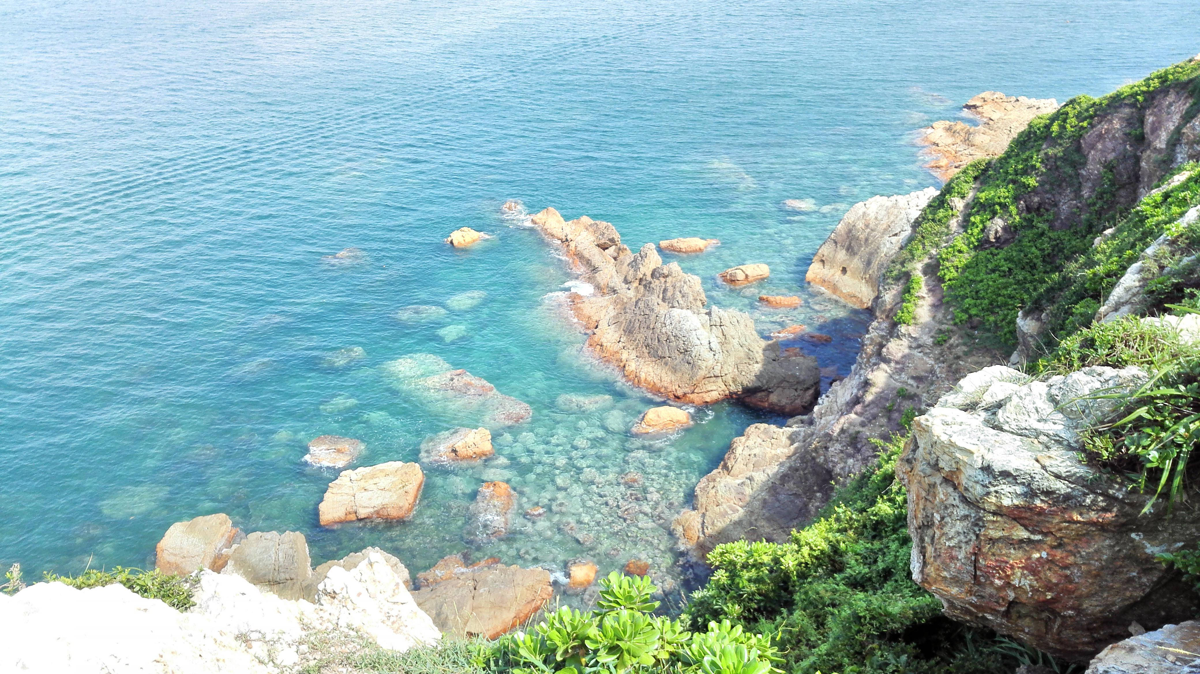 深圳大鵬半島 壁紙 — 陽光照射下的碧綠海水輕輕拍打著覆蓋著茂密綠植的崎嶇海岸岩石。