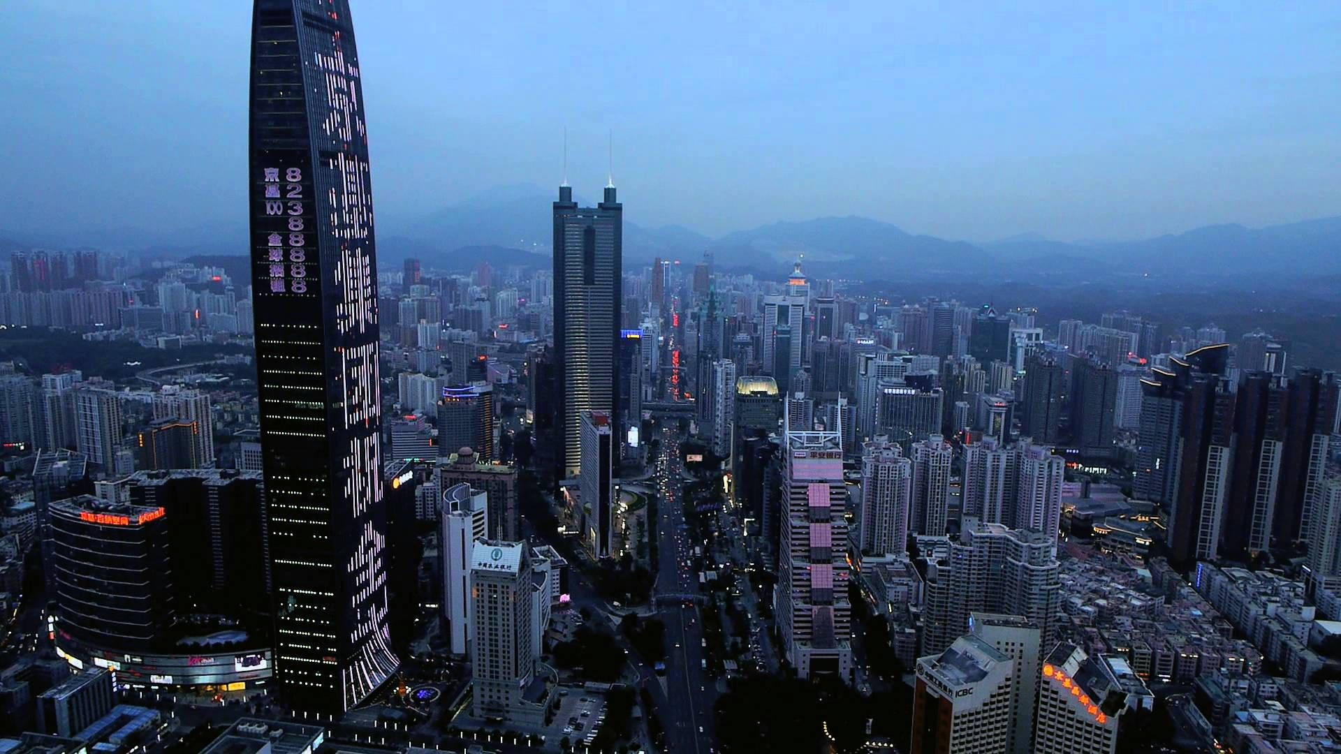 深圳霧夜城市 壁紙 — 黃昏時分廣闊城市景觀的鳥瞰圖，高聳的摩天大樓在漸暗的藍天和遠山映襯下亮起燈光。