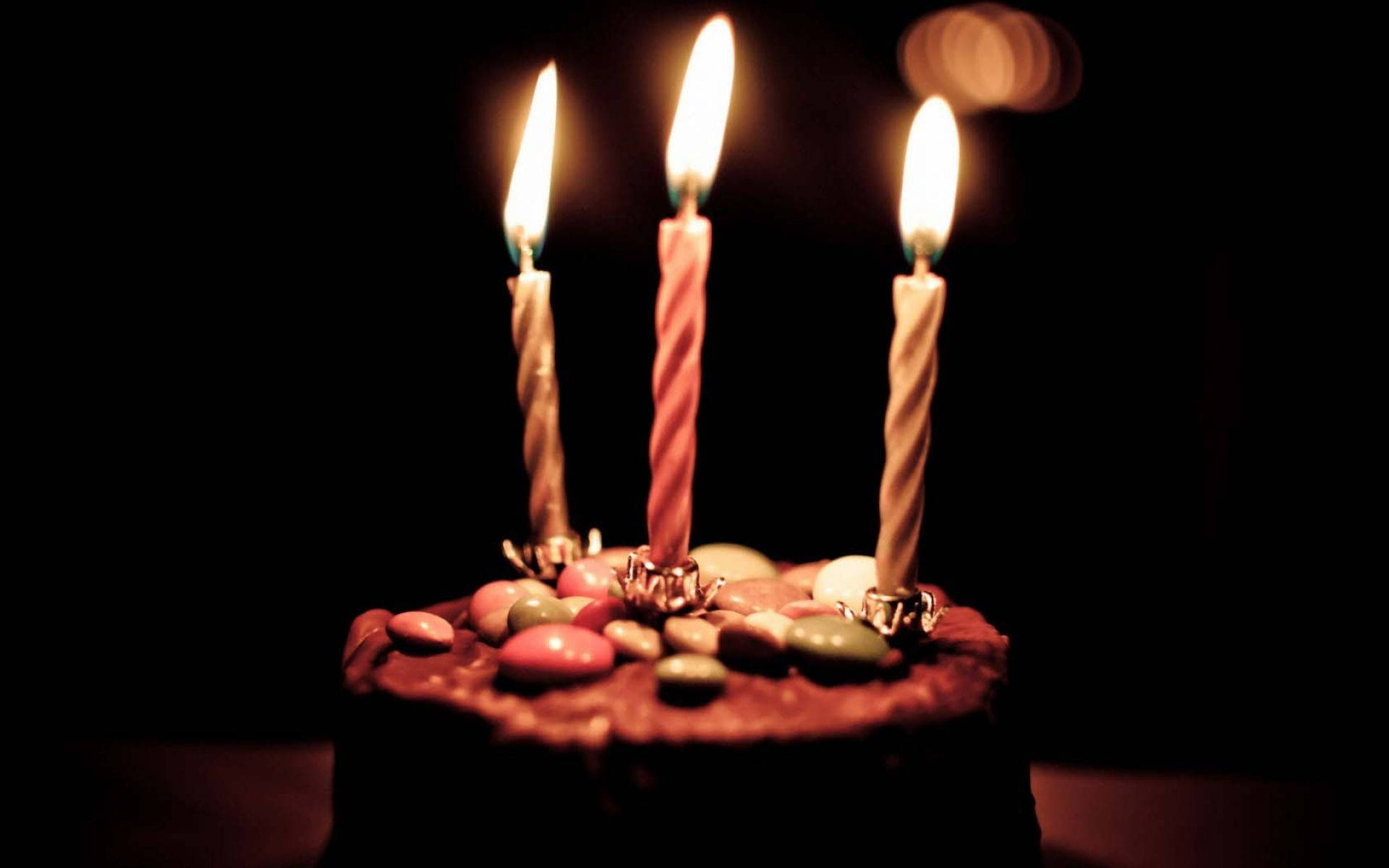 深棕色生日蛋糕蠟燭 壁紙 — 三根點燃的生日蠟燭在深色背景下的朱古力蛋糕上溫暖地發光，蛋糕上裝飾著色彩繽紛的糖果。