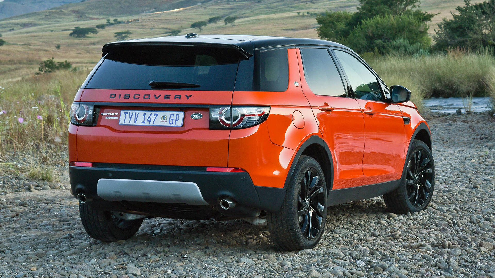 深橙色路虎 iPhone 主題壁紙 壁紙 — 一輛鮮橙色的 Land Rover Discovery Sport 停泊在碎石路上，背景是起伏的山丘和乾草。