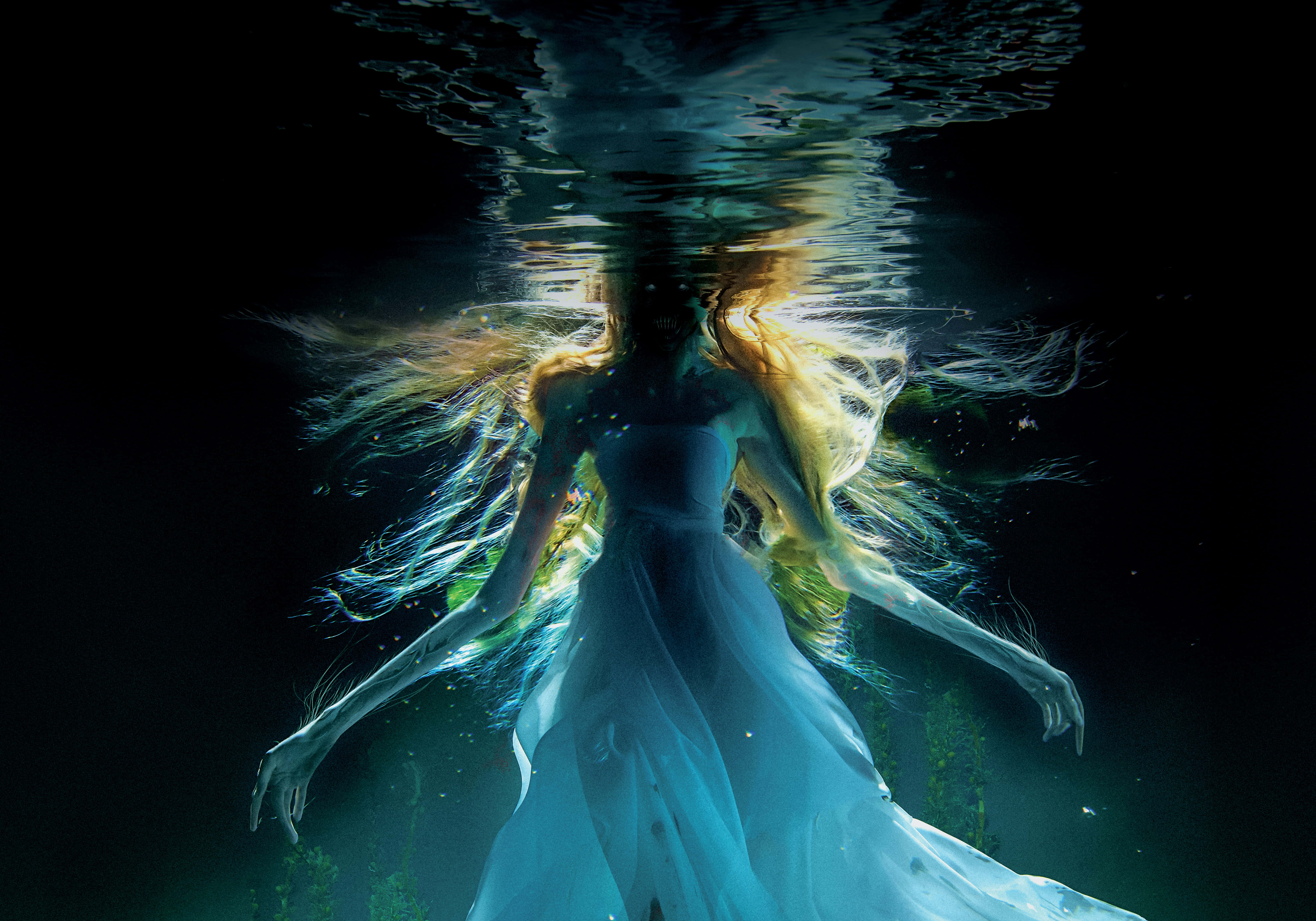 深海神秘美人魚彩色圖像 壁紙 — 一位身穿飄逸藍色長裙的女子在水中漂流，金色頭髮在她周圍盤旋，光線從上方透入。