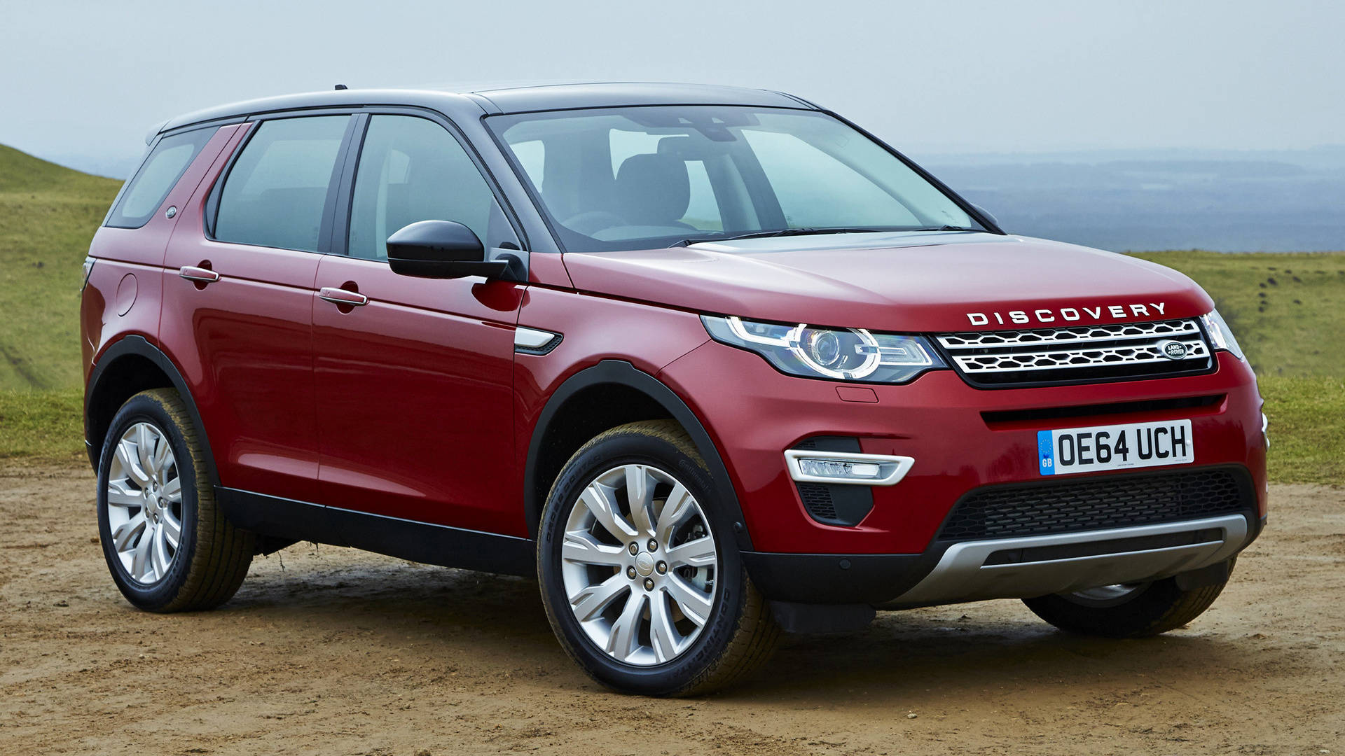 深紅色發現版路虎 iPhone 主題壁紙 壁紙 — 一架亮紅色嘅Land Rover Discovery SUV停泊喺一條泥路上，背景係起伏嘅綠色山丘，天空多雲。