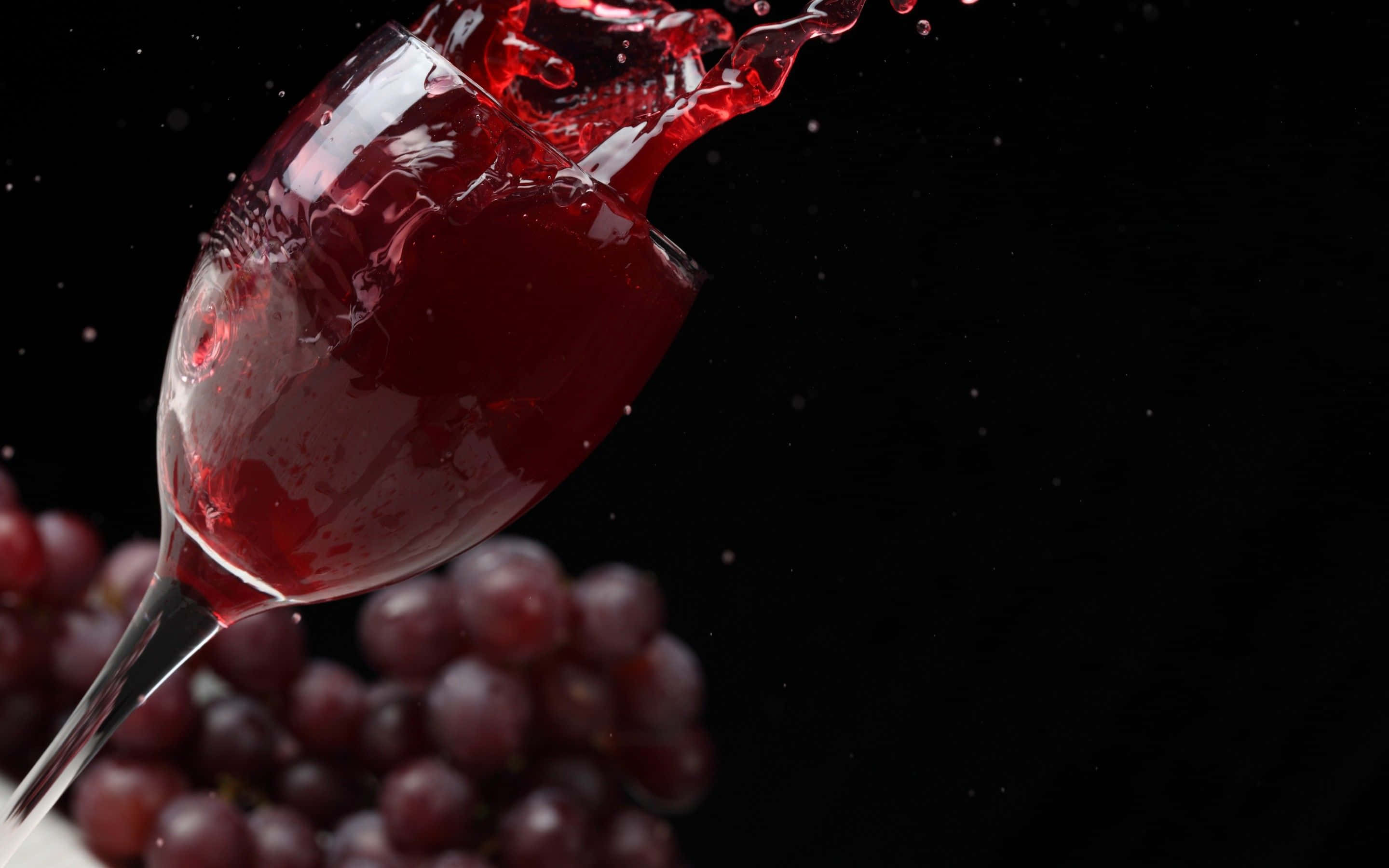 Red Wine Glass 桌布 壁紙 — 深紅酒液從傾斜的玻璃杯中動態噴濺，黑色背景下方有模糊的紫色葡萄。
