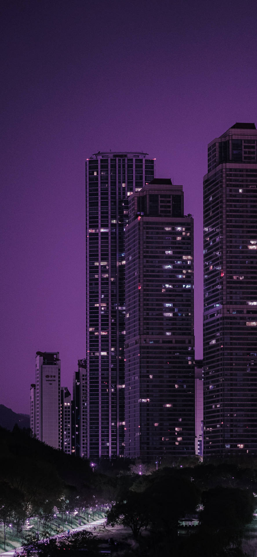 深紫色夜空下的摩天大樓 壁紙 — 高聳的摩天大樓在深紫色暮色天空下，沿著一條黑暗的林蔭大道散發著溫暖的光芒。