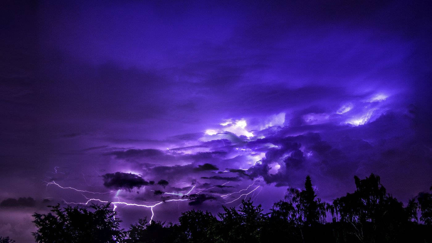 紫色閃電劃過暴風雲層，照亮夜間黑暗的樹木剪影。