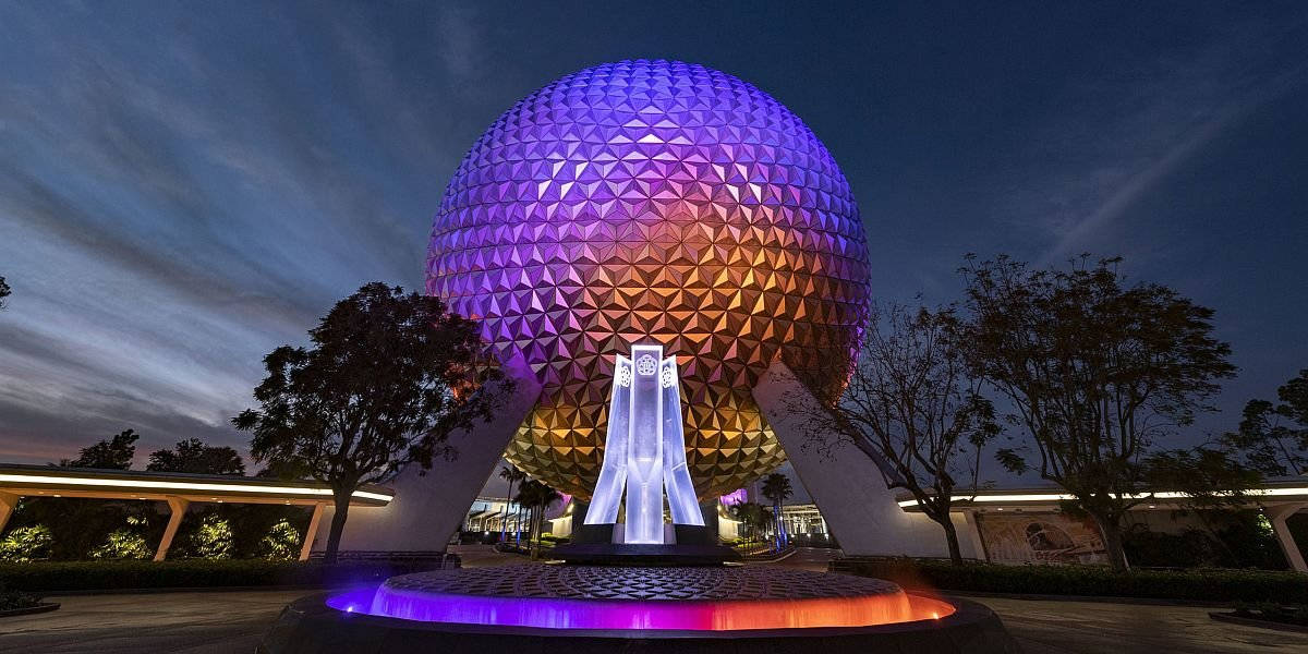 深色天空下的色彩繽紛 Epcot 球體 壁紙 — 一個巨大的測地線球體在暮色天空下，在未來主義噴泉上方發出紫色和橙色的光芒。