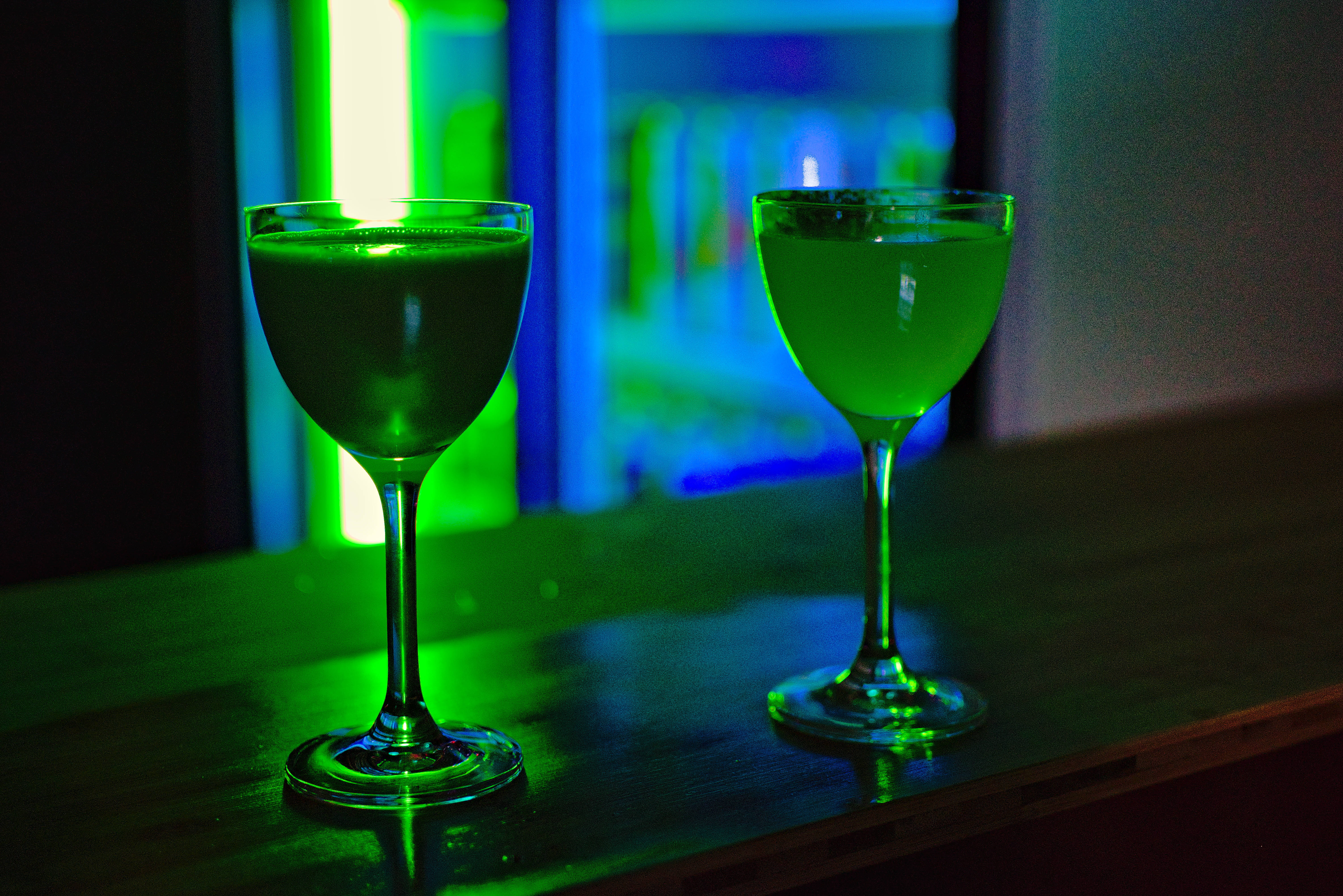 深色少女風飲品背景 壁紙 — 兩杯發光的綠色雞尾酒盛於高腳杯中，放置在黑暗的吧檯上，霓虹燈光投射出鮮豔色彩。