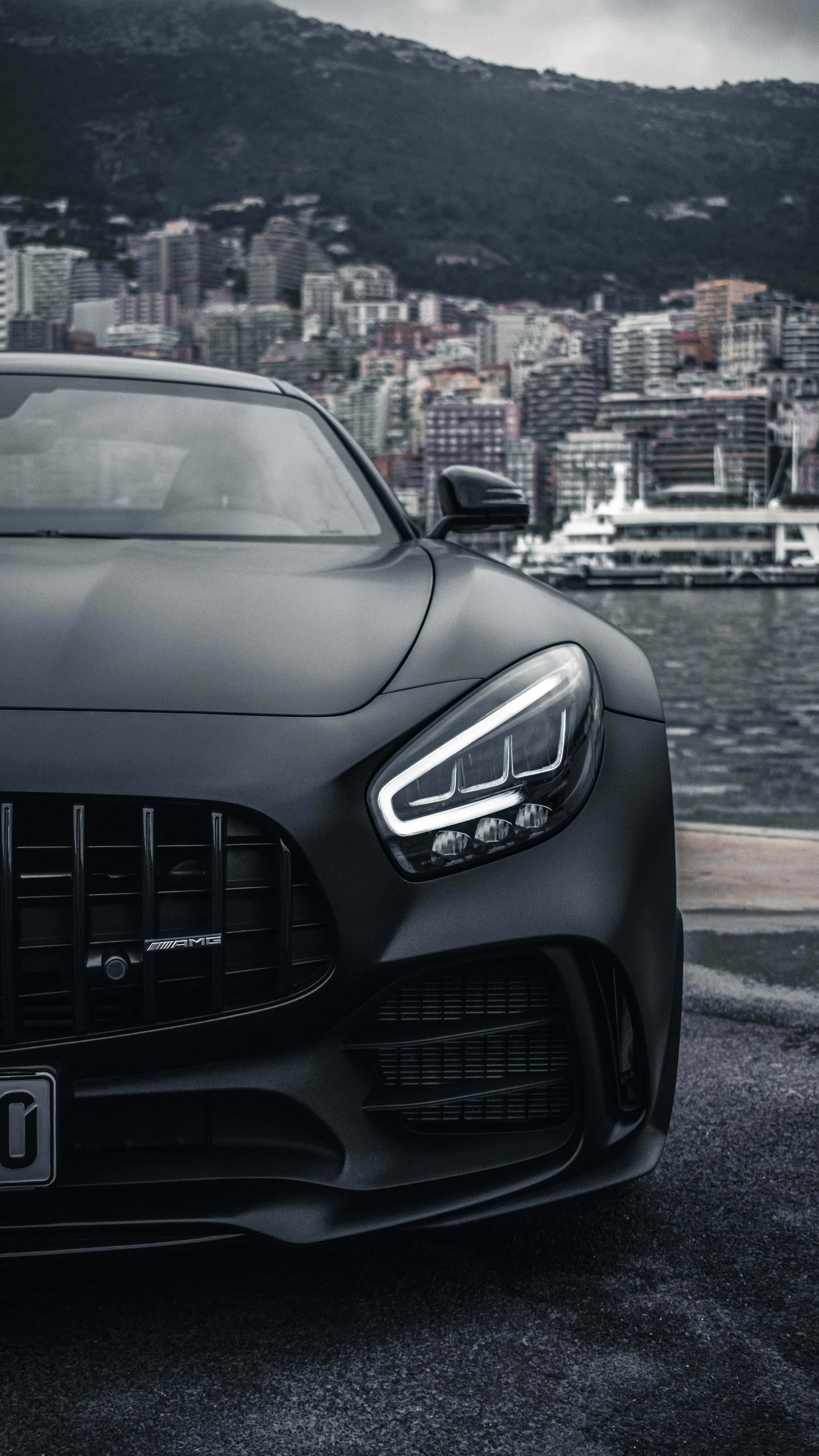 深色梅賽德斯-AMG GT R 背景 壁紙 — 一架啞黑色AMG跑車停泊在海濱，背景係城市景觀同山脈，天色陰沉。
