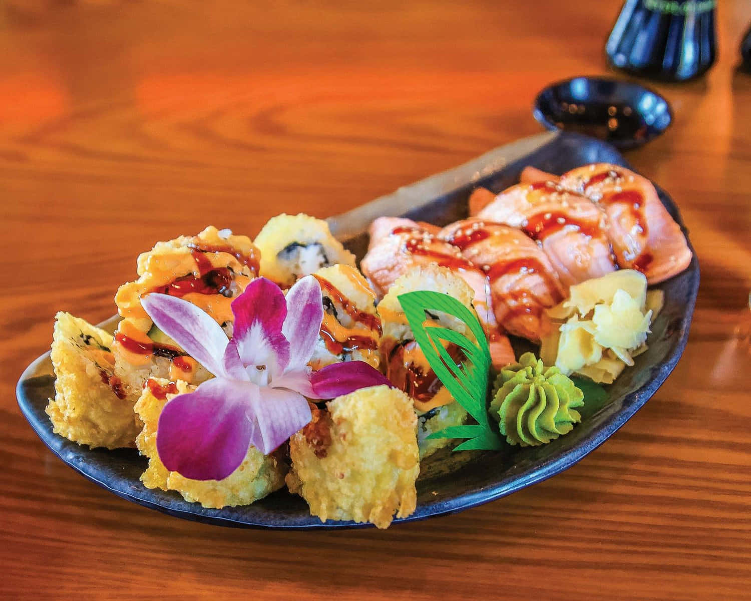 Sushi 桌布 壁紙 — 深色碟子上放著金黃色的天婦羅卷和釉面三文魚刺身，飾以紫色蘭花和芥末，放在溫暖的木桌上。