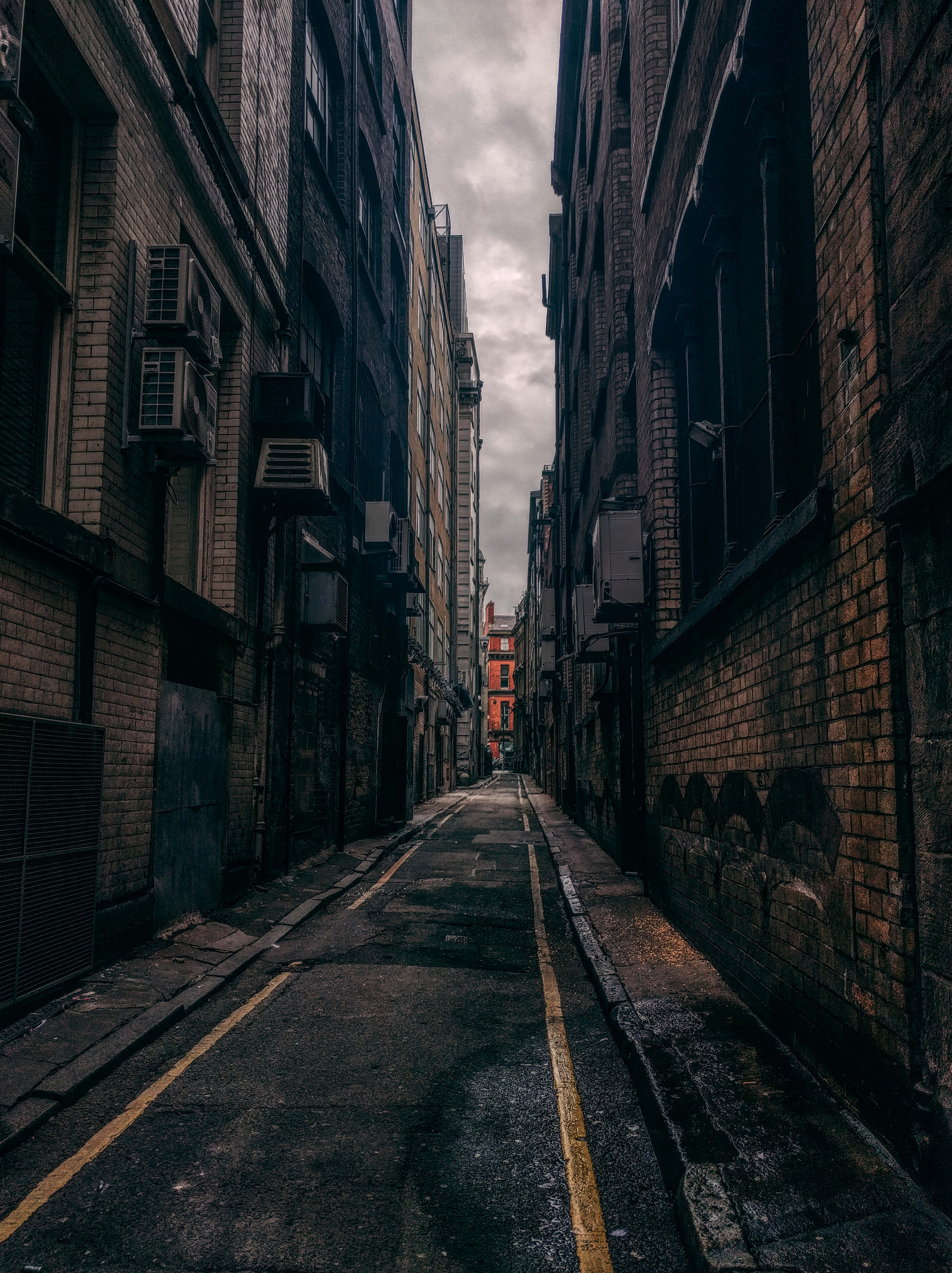 深色磚牆建築巷弄背景 壁紙 — 高樓磚建築之間狹窄的城市小巷，陰天，黃色道路標線。