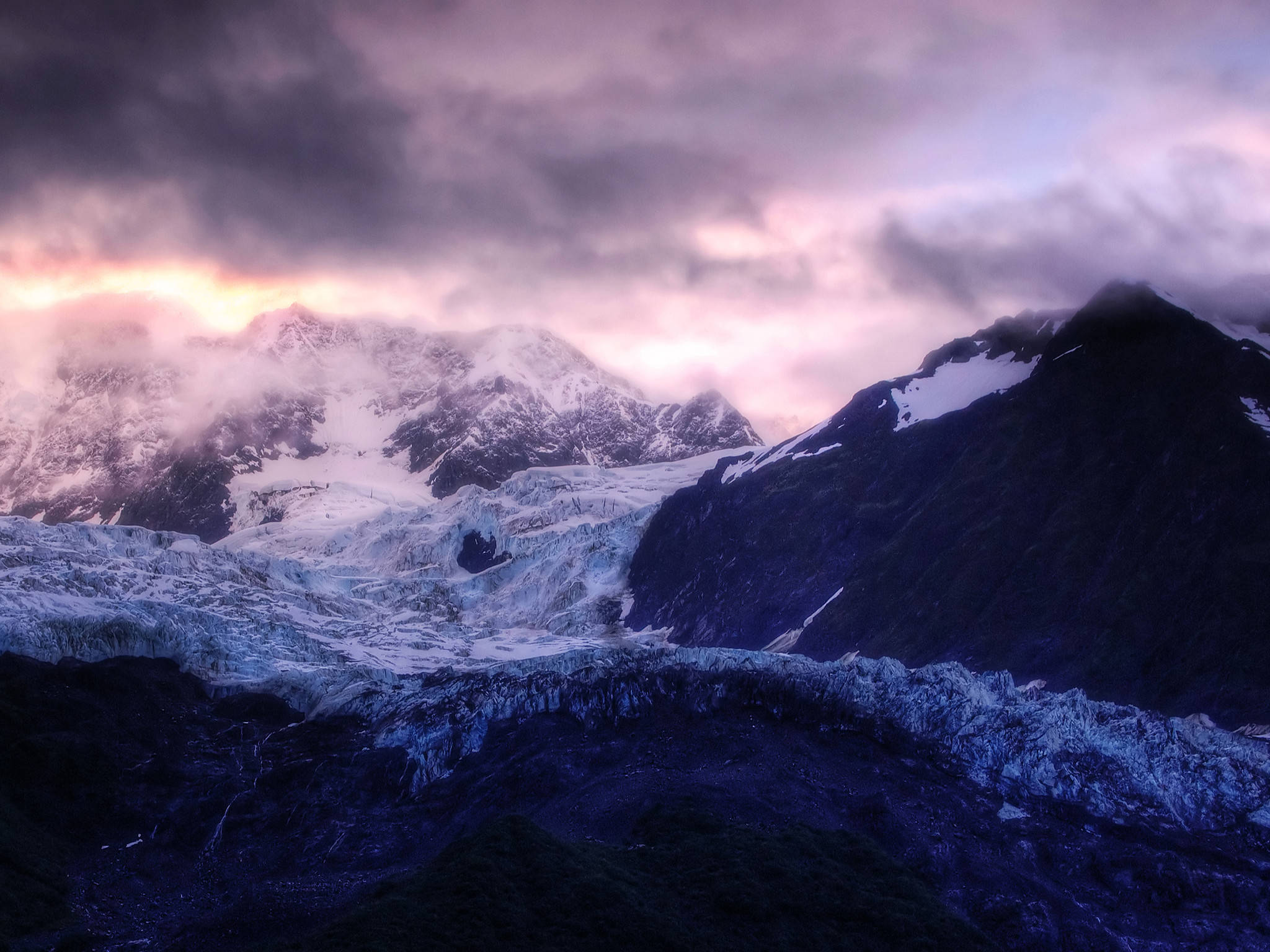 深藍海洋靜電圖 壁紙 — 一條宏偉的冰川蜿蜒穿過積雪的山峰，在戲劇性的紫色和粉紅色暮色天空下。