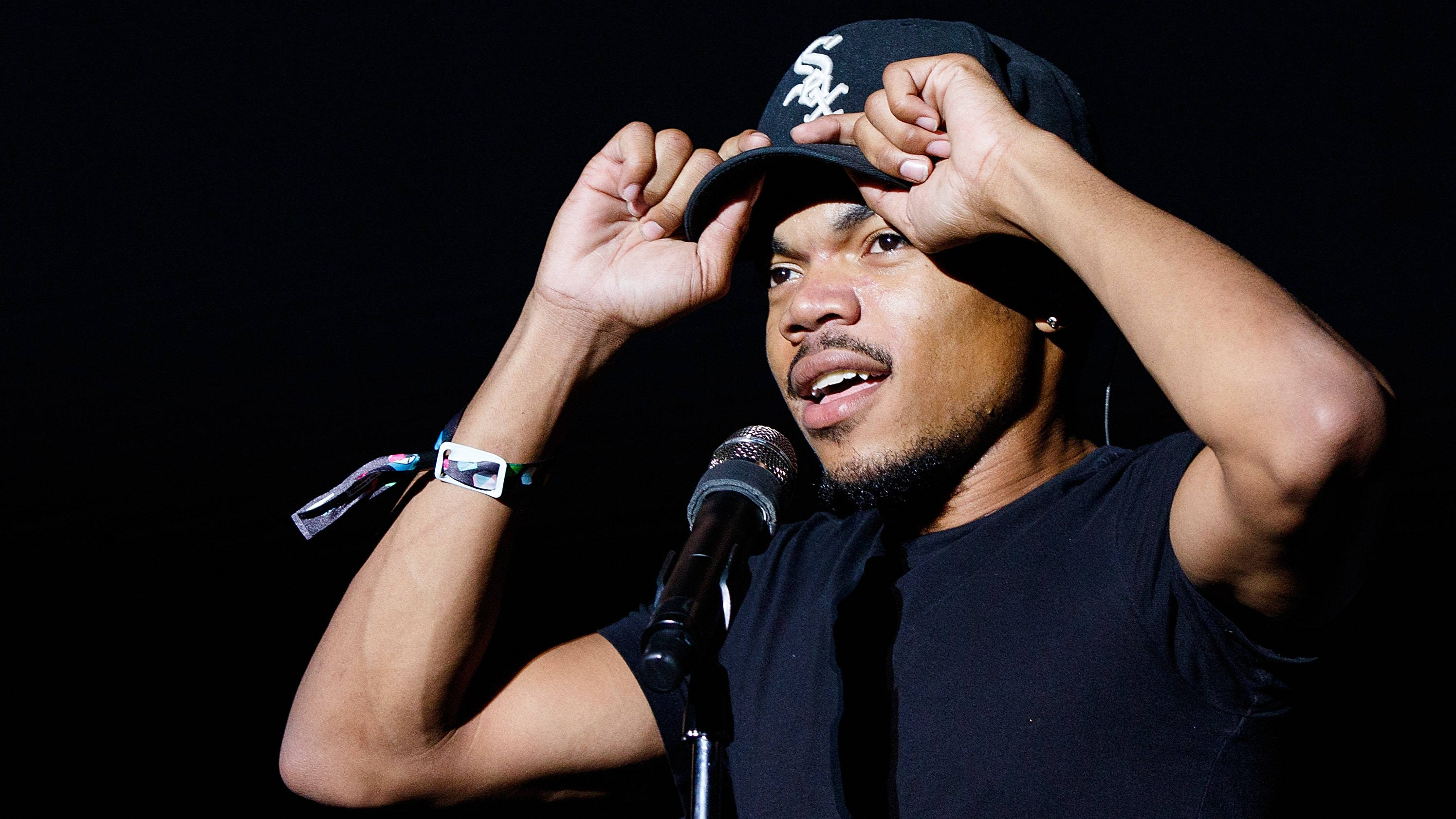 深藍色的Chance the Rapper 壁紙 — 一名戴黑色鴨舌帽的男子在舞台燈光下調整帽子，同時對著麥克風唱歌。