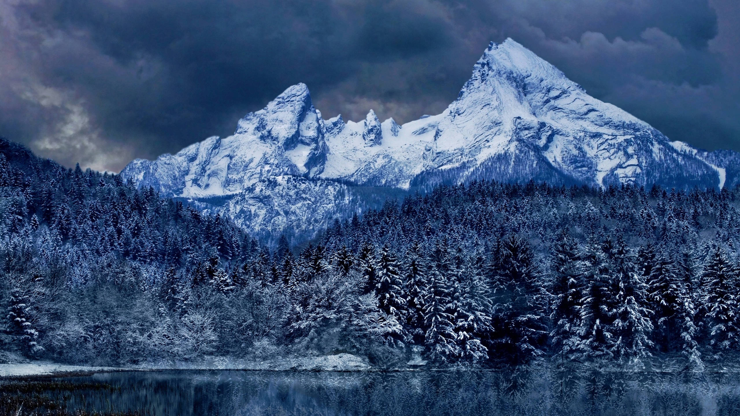 深藍色雲層籠罩山脈 壁紙 — 積雪的山峰在風暴天空下聳立，倒映在平靜湖泊中的冰凍森林之上。