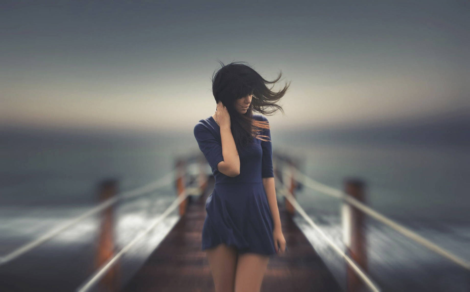 身穿藍色連衣裙的女性在黃昏時站在碼頭上，風吹動她的頭髮。