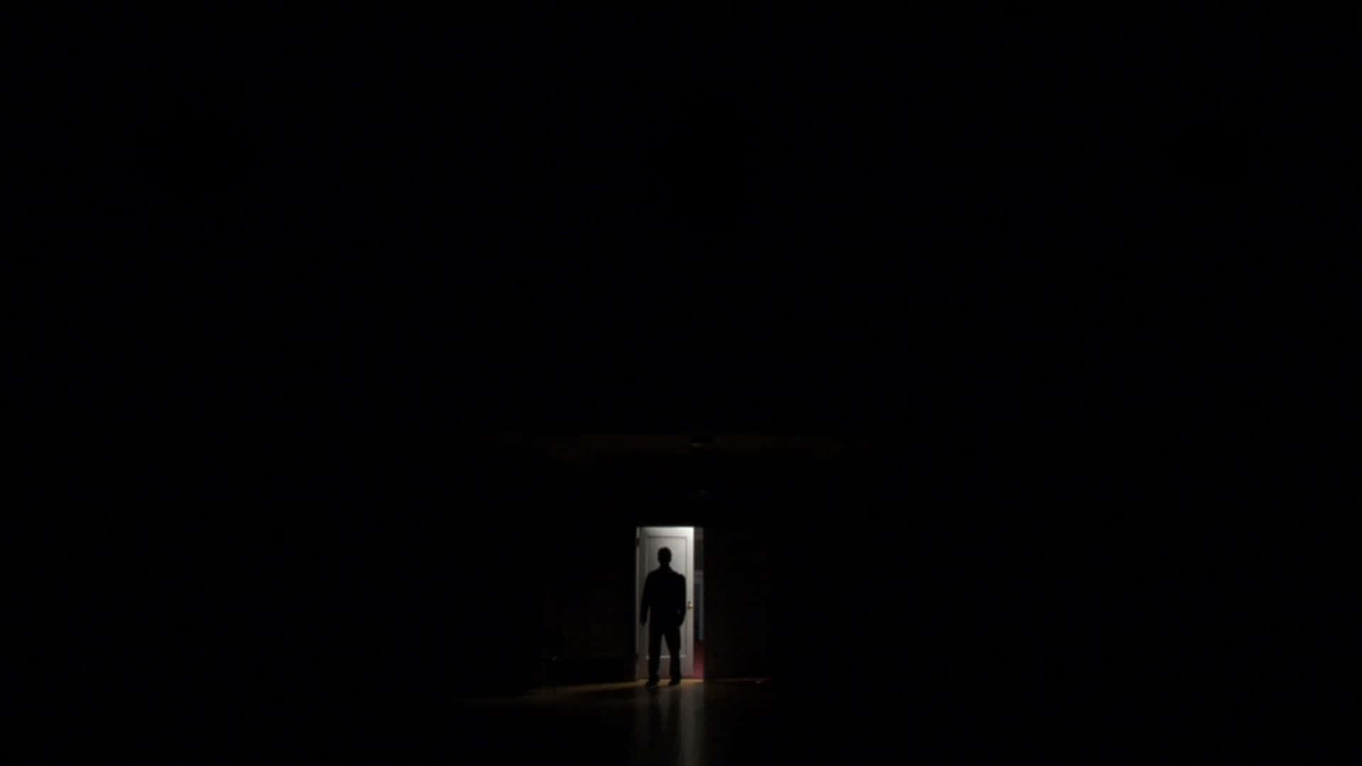 一個人站在黑暗走廊盡頭的門口，在明亮光線下形成剪影。