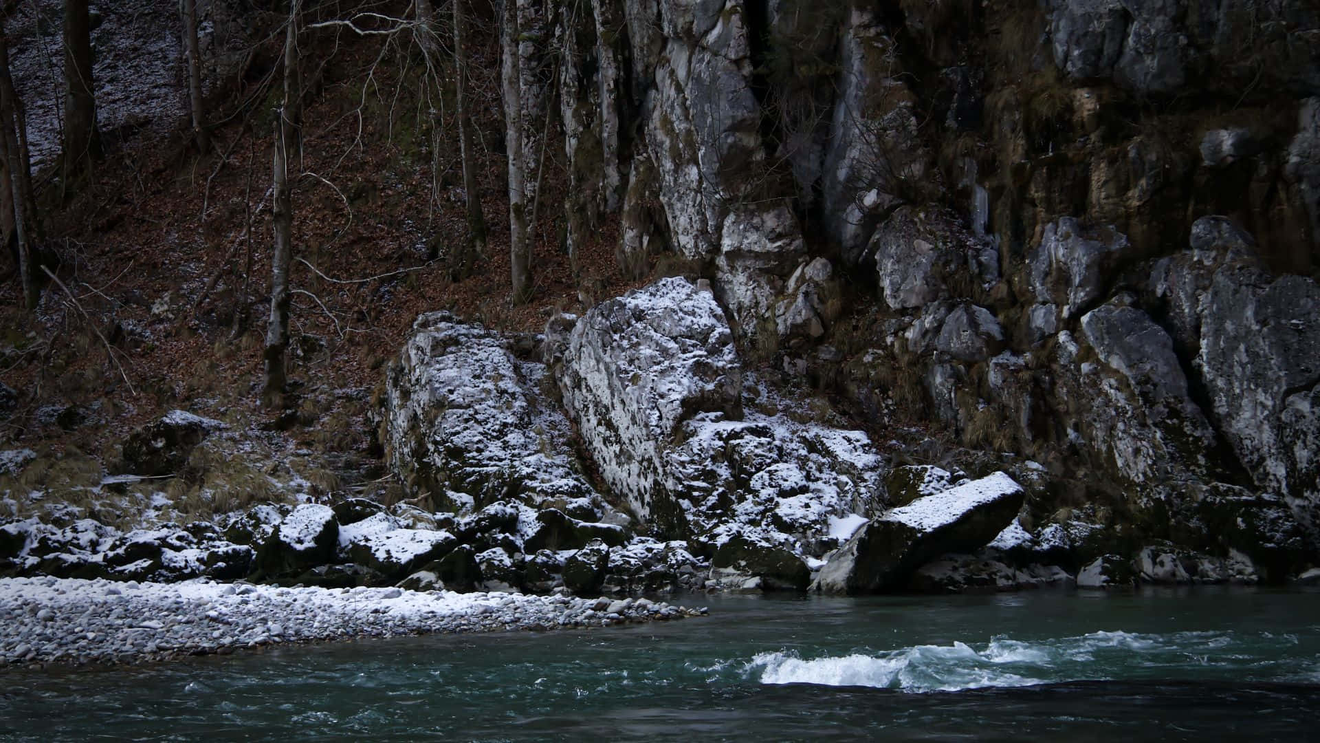 一條積雪的河流流過岩石河岸和光禿禿的樹木，冬季景象。