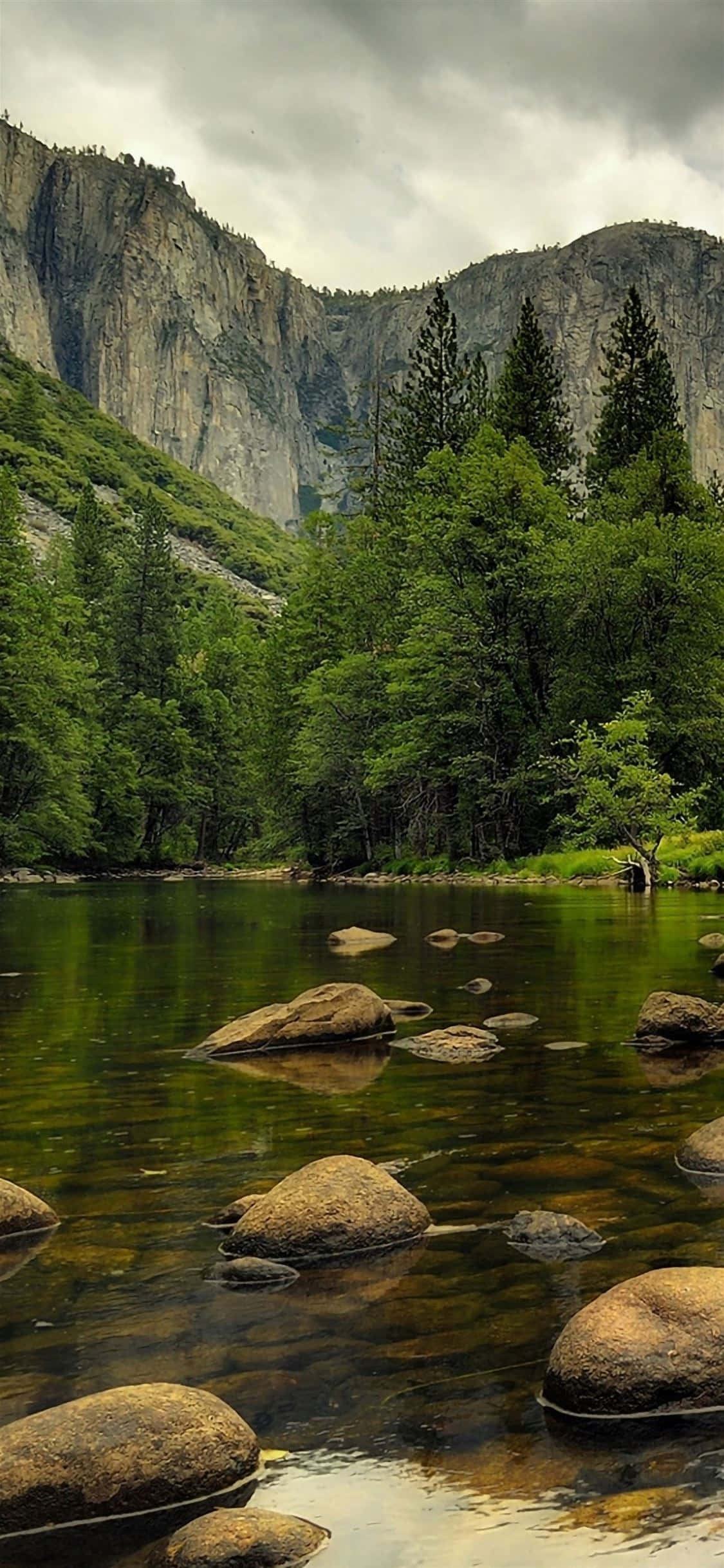 淺溪自然風景 壁紙 — 一條寧靜的河流流過長滿苔蘚的岩石，周圍是茂密的綠色森林和聳立的花崗岩懸崖，天空多雲。