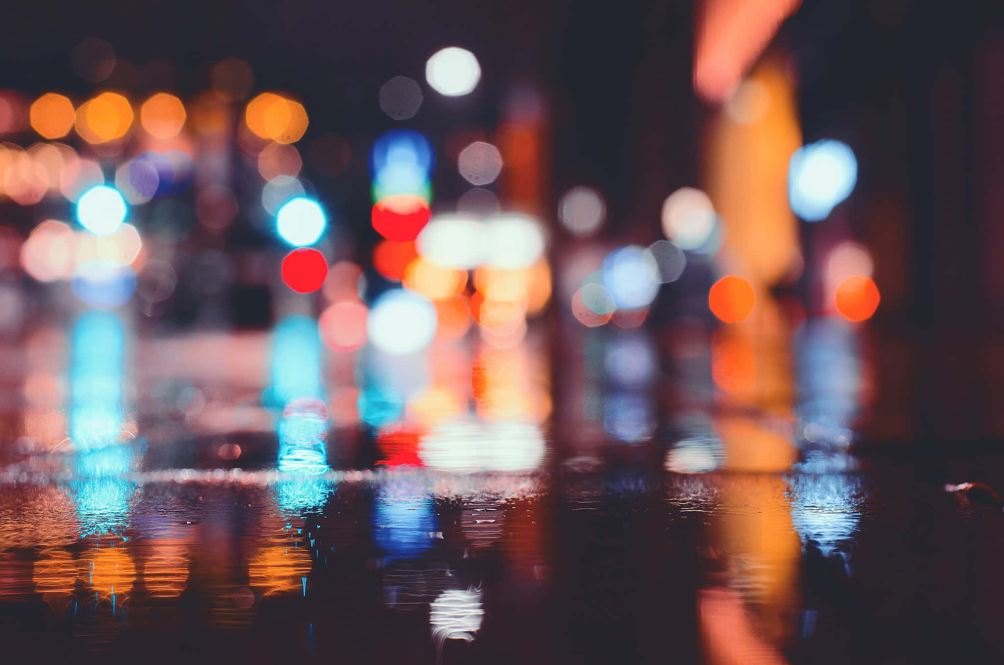 淺色模糊城市燈光圖案 壁紙 — 雨濕的夜間城市街道，鮮豔的散景燈光在水坑中反射，營造出夢幻般的發光效果。
