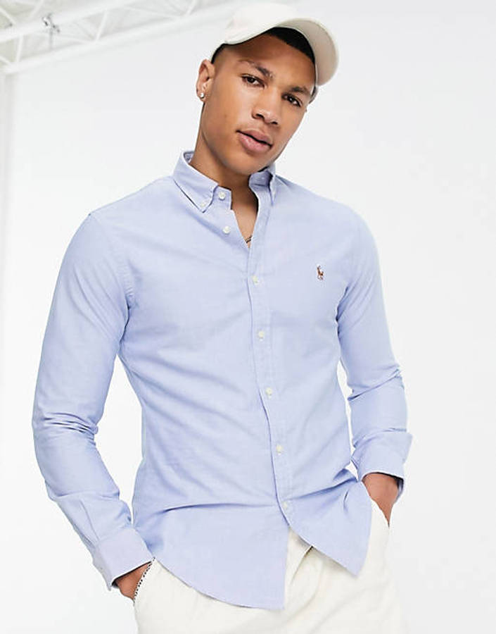 淺藍 Ralph Lauren 按鈕開襟Polo衫 壁紙 — 一位年輕男子身穿淺藍色鈕扣襯衫和白色帽子，雙手插袋，在明亮的攝影棚背景下自信地擺姿勢。