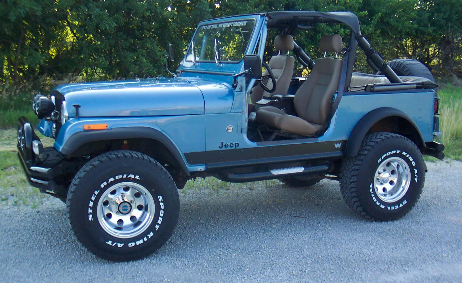 Jeep Cj 桌布 壁紙 — 淺藍色Jeep Wrangler，配備米色內飾及大型越野輪胎，停泊在碎石路上，附近有綠樹。