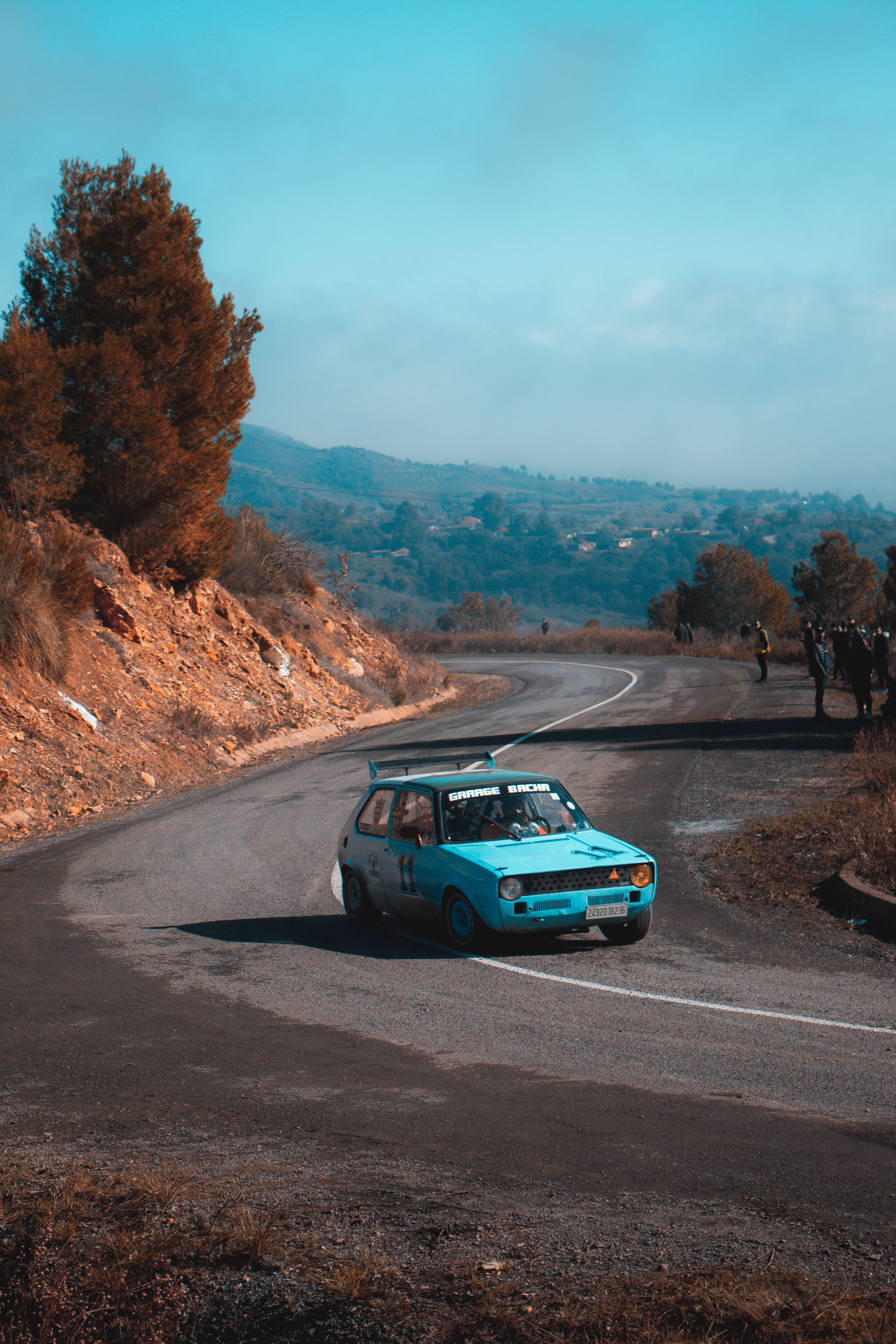 淺藍色賽車在 Dirt Rally 遊戲中競速 壁紙 — 一輛充滿活力的青綠色拉力賽車沿著蜿蜒的山路飛馳，觀眾在朦朧的藍天下從路邊觀看。