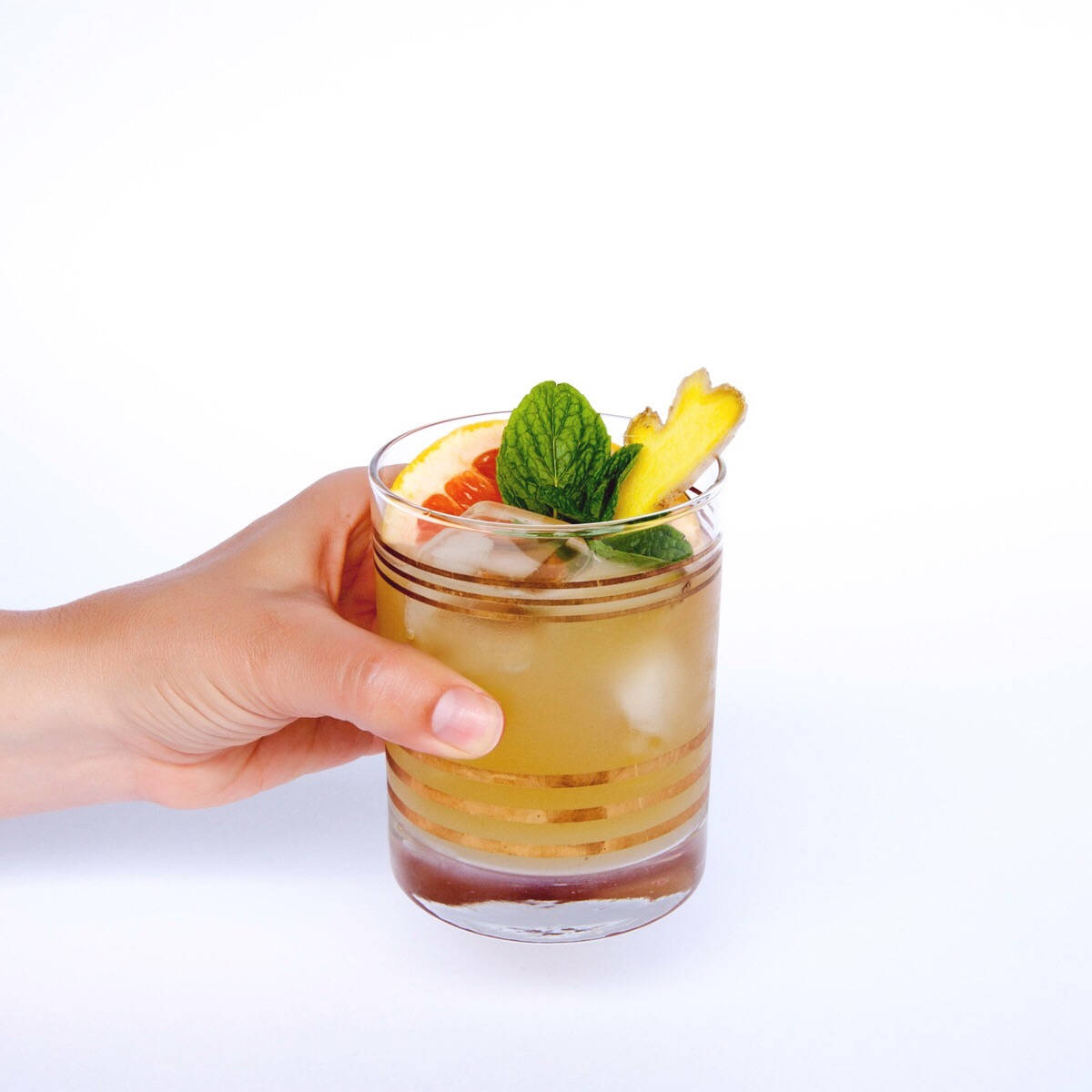 清新水果糖漿飲品 壁紙 — 一隻手拿著金色條紋玻璃杯，杯內盛載琥珀色雞尾酒，飾以薄荷、西柚和菠蘿片，背景為白色。