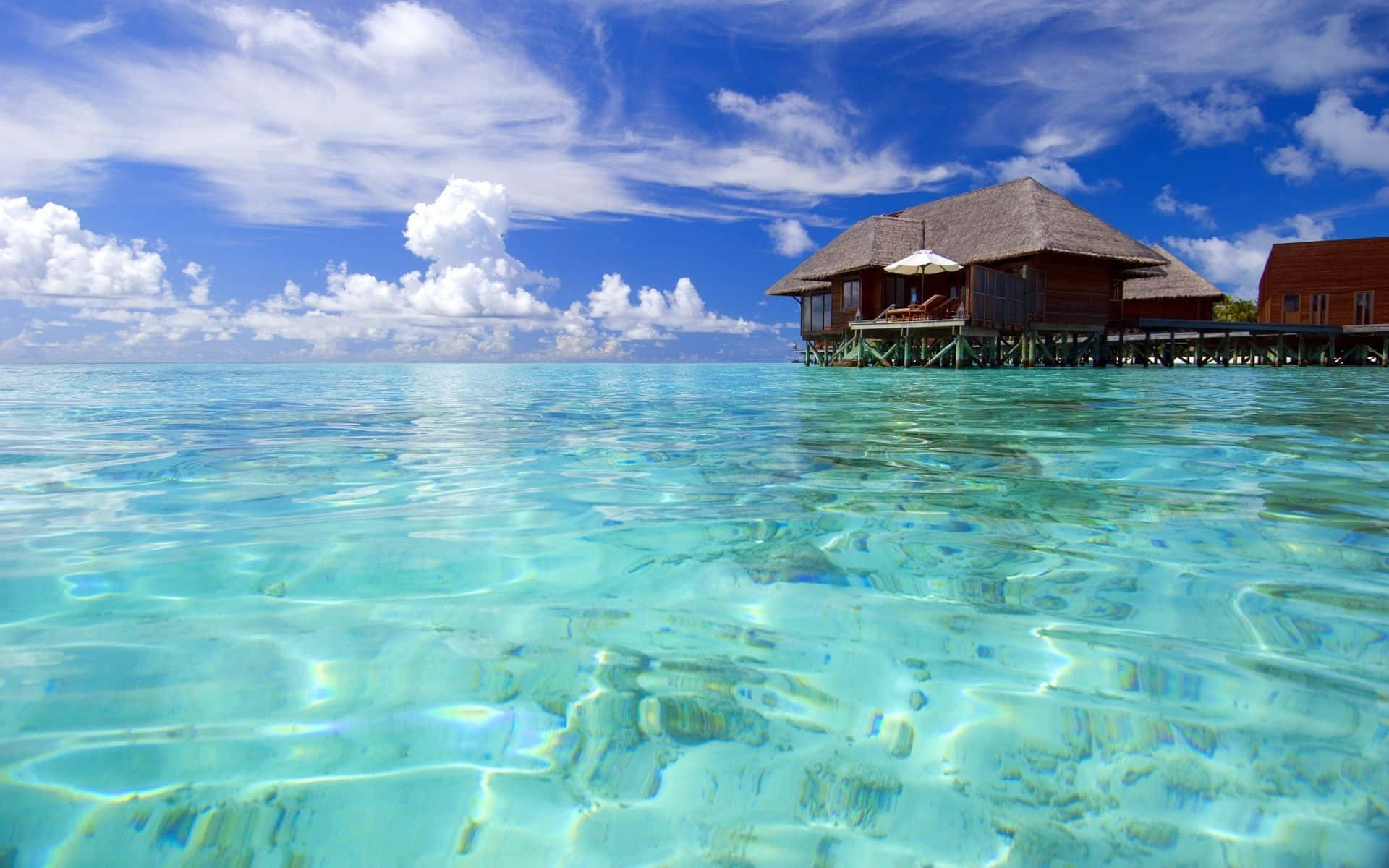 Maldives Beach 桌布 壁紙 — 清澈碧綠的海水在木製水上平房周圍泛起漣漪，上方是明亮的藍天和蓬鬆的白雲。