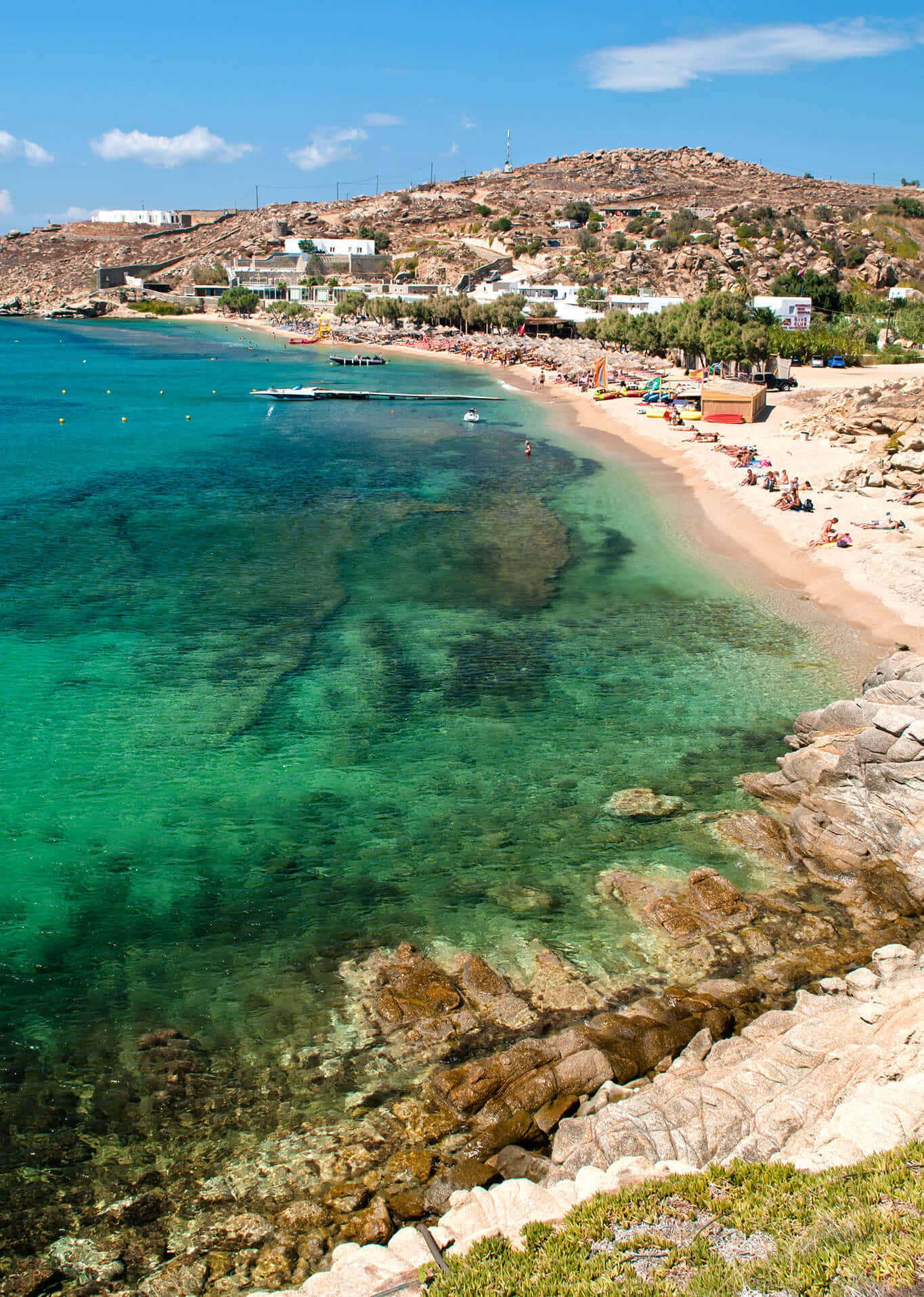 Greek Beach 桌布 壁紙 — 清澈碧綠的海水拍打著岩石海岸，海灘遊客在明亮的藍天下放鬆身心。